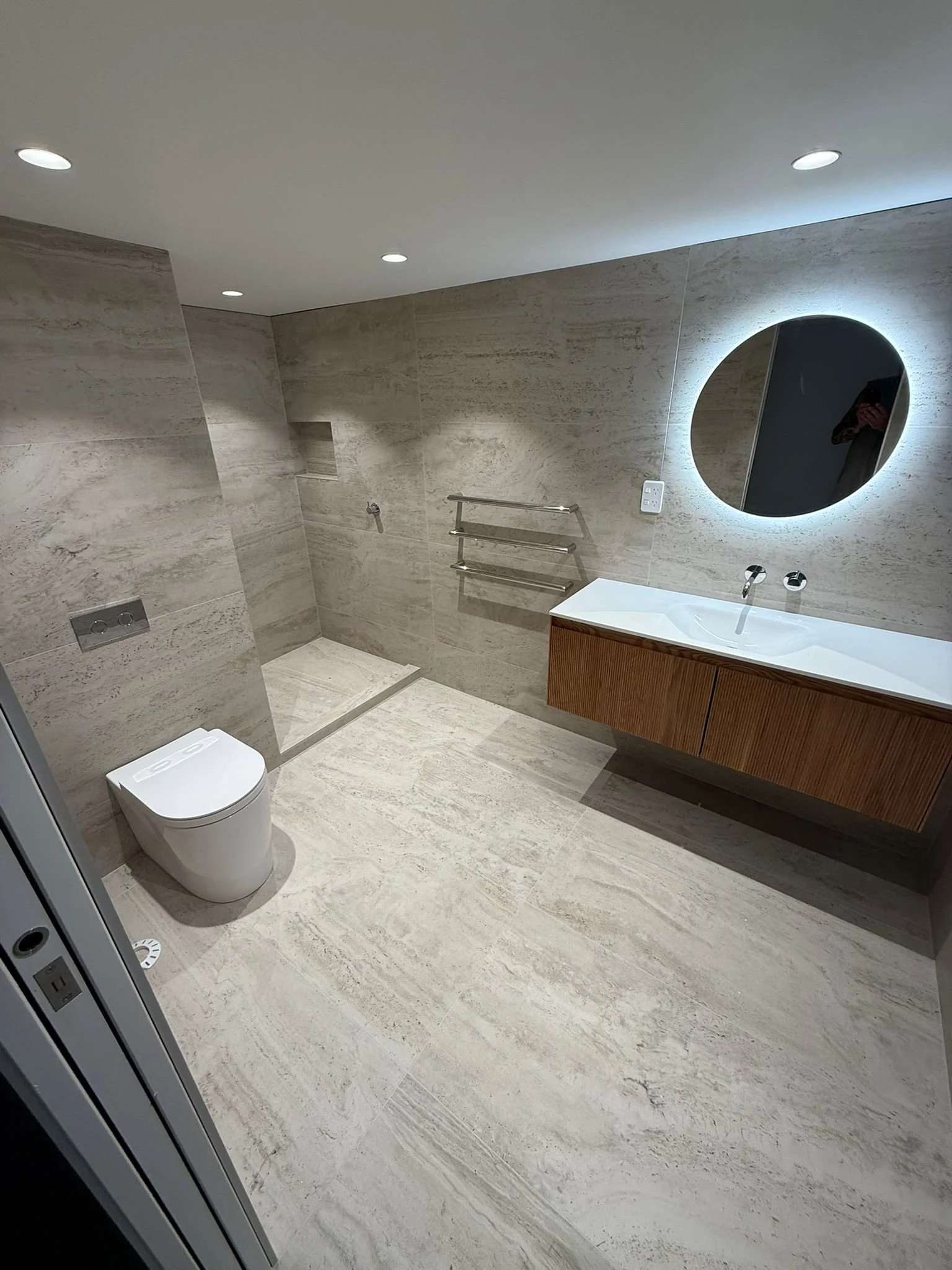 Modern bathroom with beige stone tiles, a wall-mounted toilet, a walk-in shower with a niche, a wooden vanity with a white sink, a round backlit mirror, and chrome fixtures.