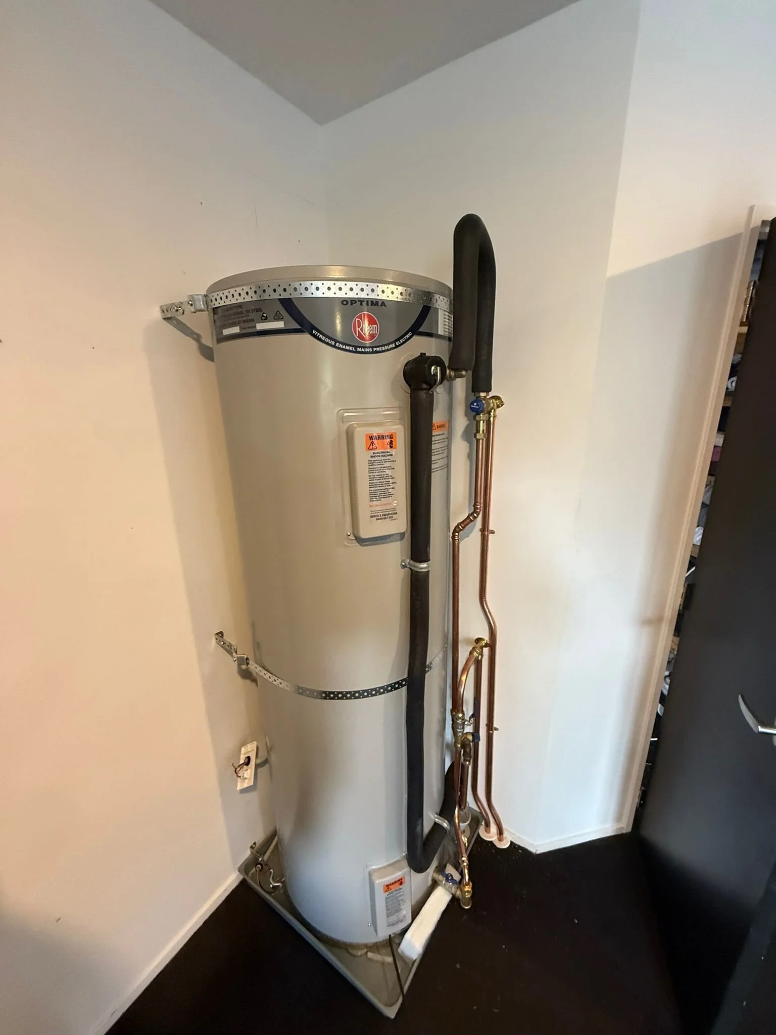 A water heater with black and copper pipes attached, placed in a corner of a room with white walls and a dark floor.