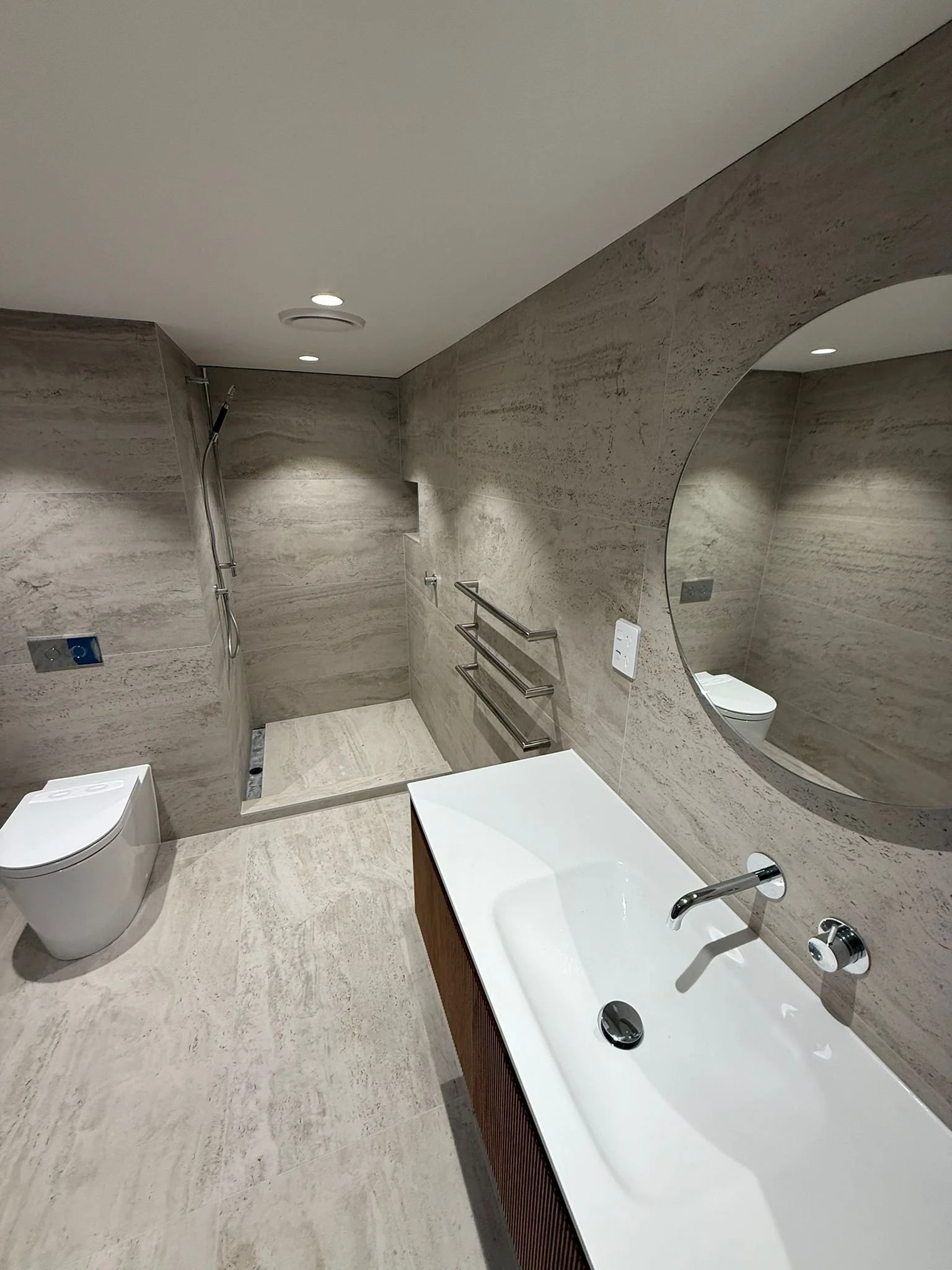 Modern bathroom with beige tile walls and floors, a walk-in shower with a handheld showerhead, a white toilet, and a white sink with a wooden vanity. A large round mirror is above the sink, and there are recessed ceiling lights.
