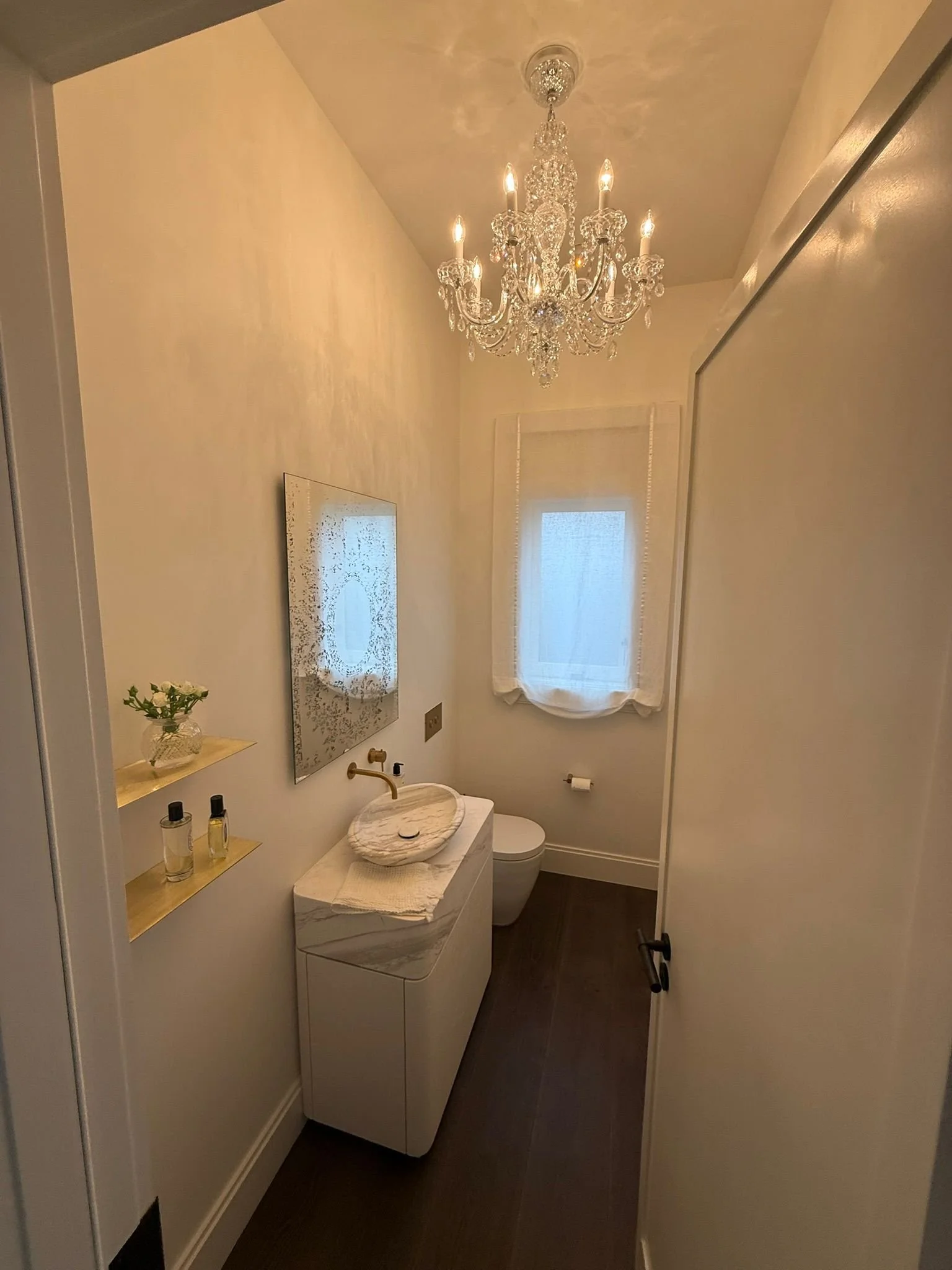 A small elegant bathroom with a crystal chandelier hanging from the ceiling, a window with sheer curtains, a white modern vanity with a marble sink, a mirror, and some decorative bottles and a small plant on a wall shelf.