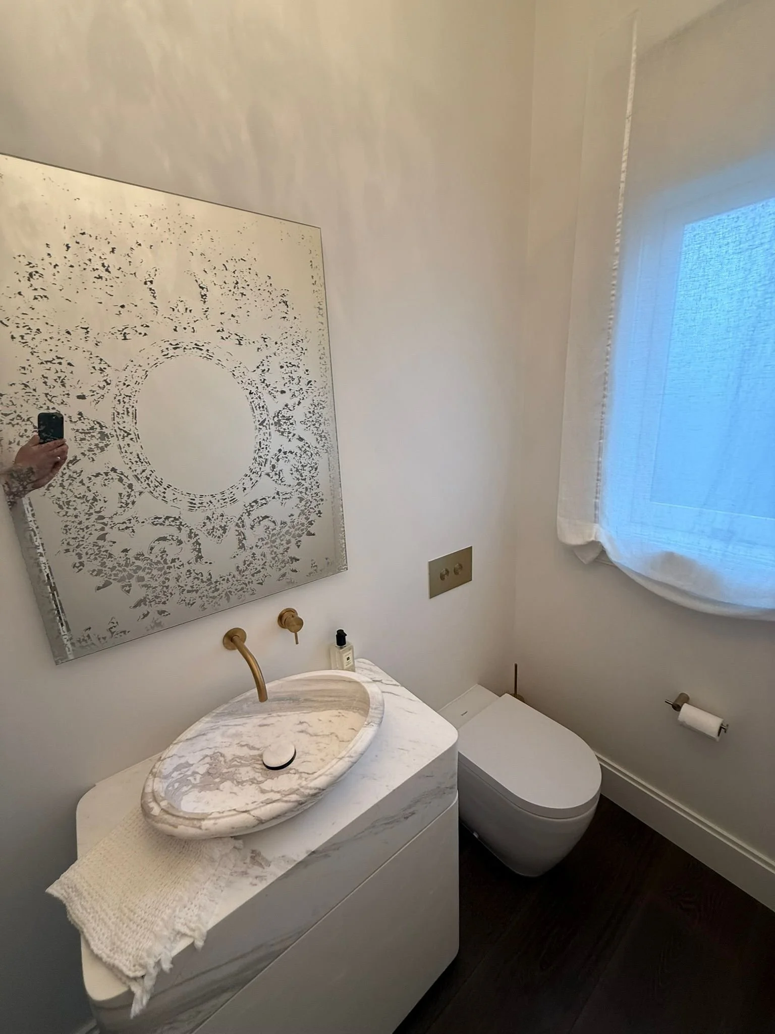A small bathroom with a white marble sink, a gold faucet, a mirror with a decorative pattern, a white toilet, a window with sheer curtains, and a dark wooden floor.