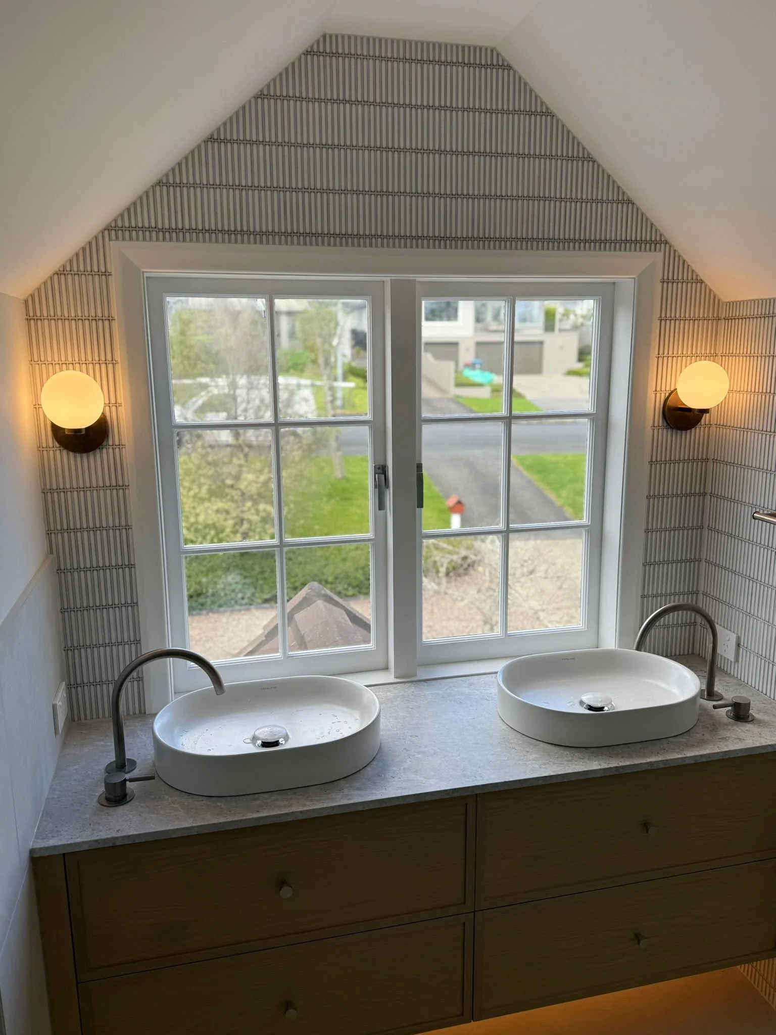 Bathroom with two oval sinks on a marble countertop, wooden vanity below, large window overlooking a green yard, two wall-mounted spherical lights, tiled wall, and a sloped ceiling.