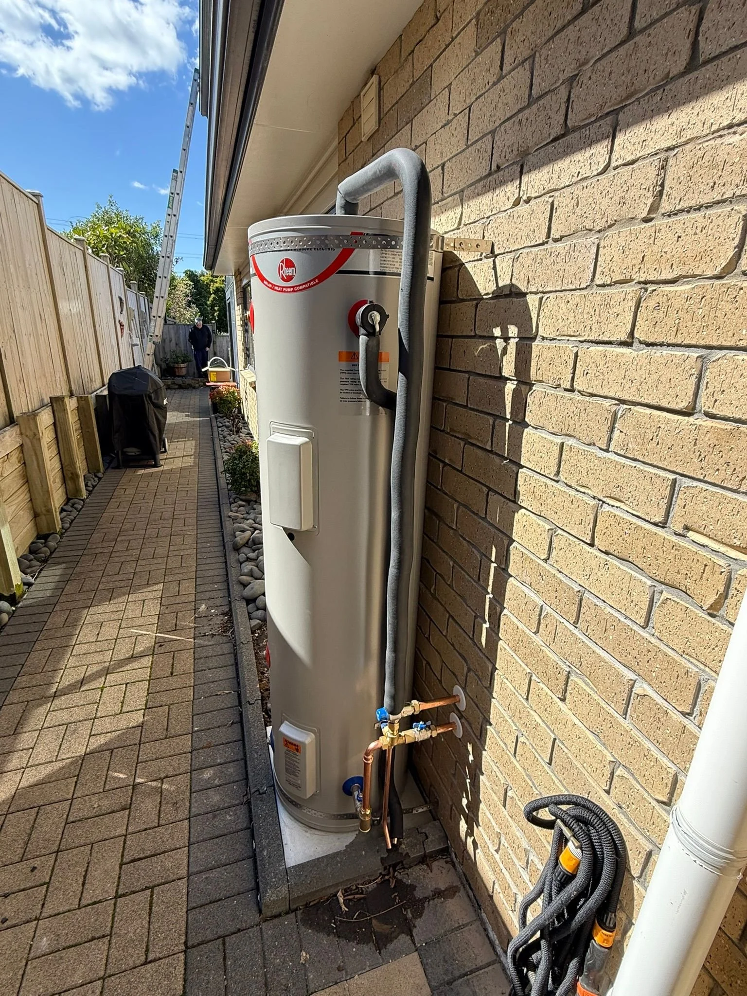 An outdoor hot water heater installed against a brick wall. Pipes and electrical cables are connected to the unit, which is positioned on a concrete base in a backyard with a paved walkway, fencing, and a person walking in the distance.