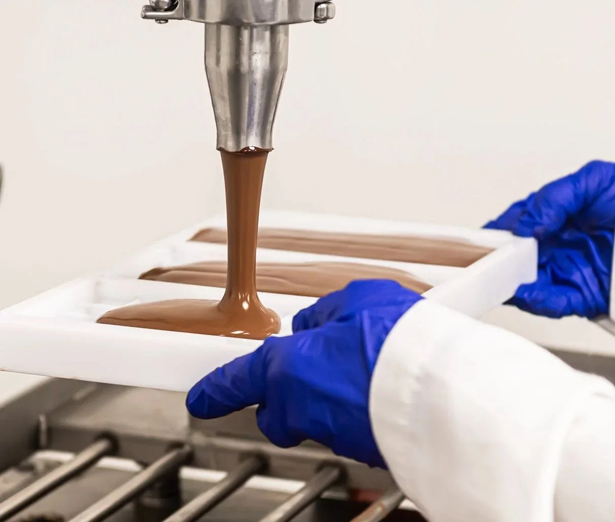 A person wearing blue gloves pours melted chocolate into a mold for making chocolates.