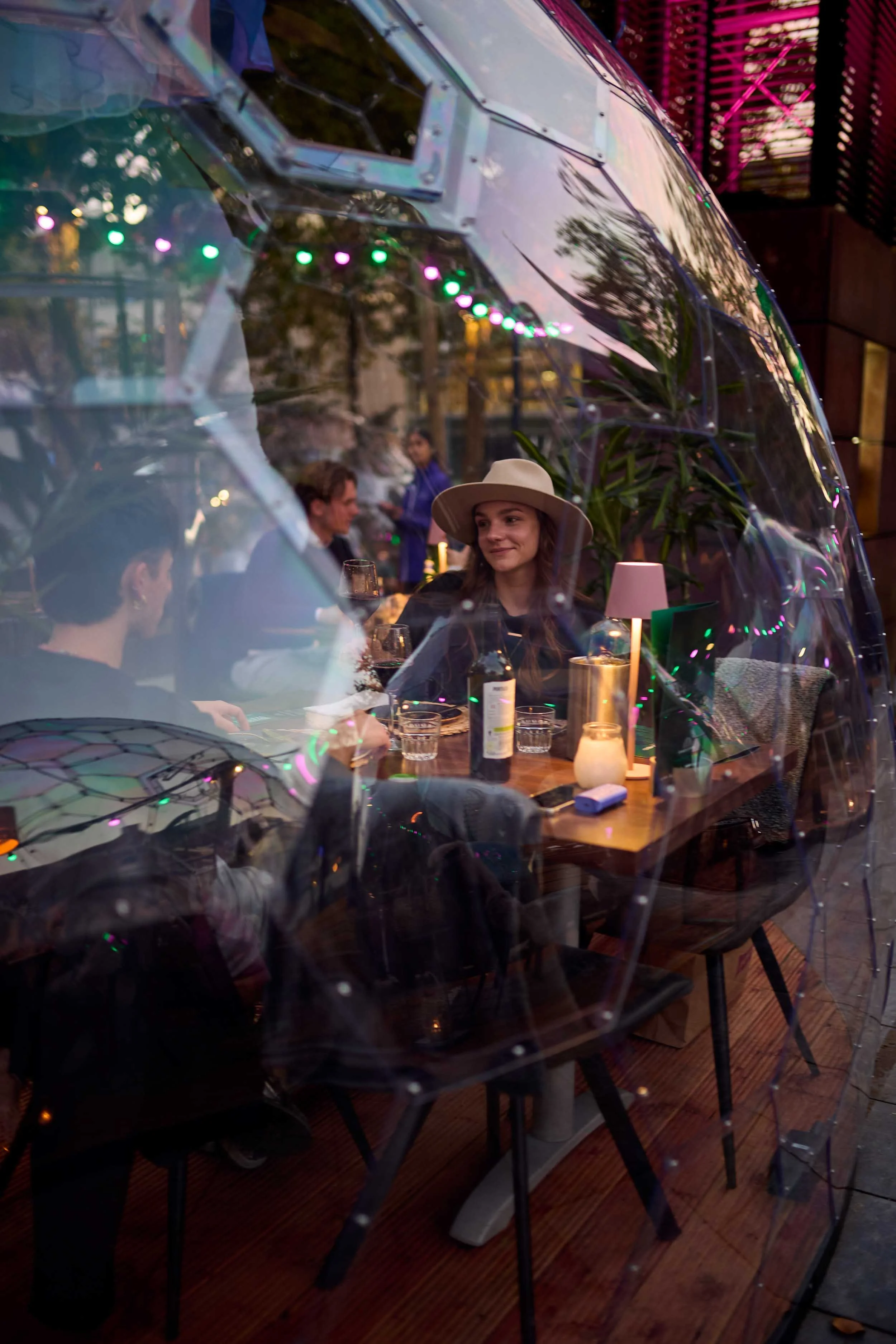 Een groep mensen zit binnen een restaurant of café, gezien door een doorzichtige glazen koepel met gekleurde lichtjes.