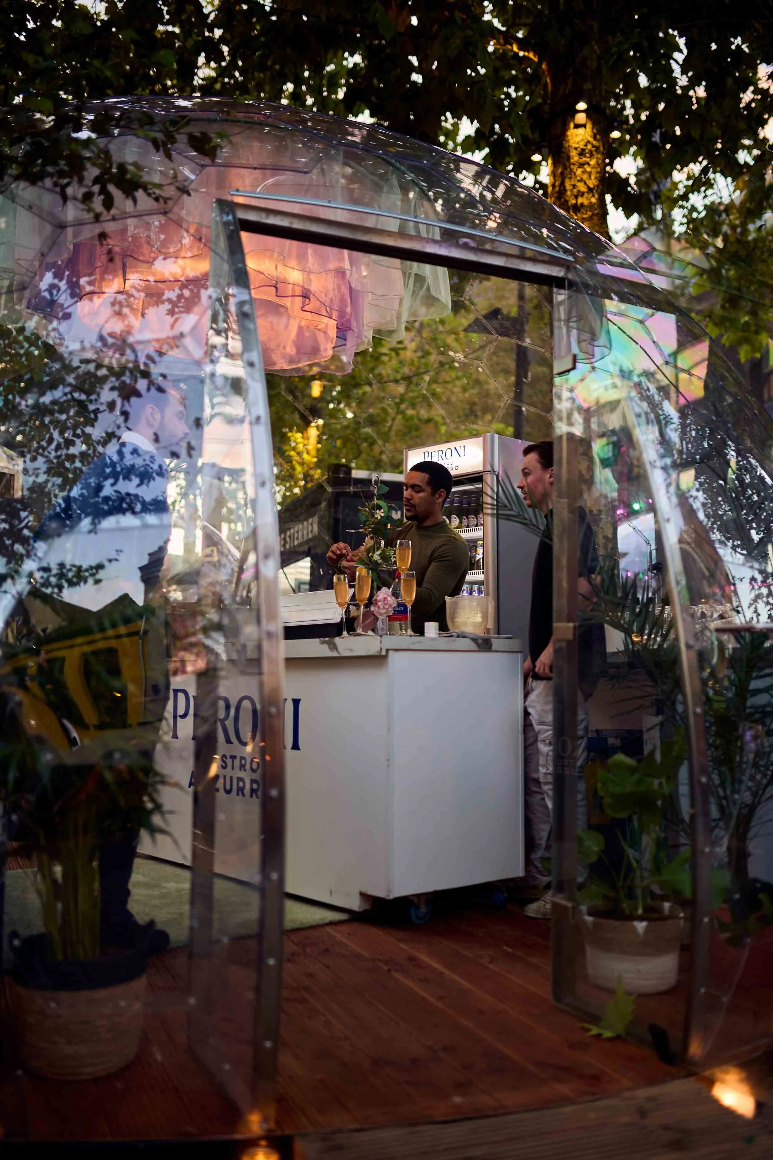 Een foto van een bar buiten onder een glazen kap, met twee barmannen die drankjes serveren, omringd door planten en bloemen.