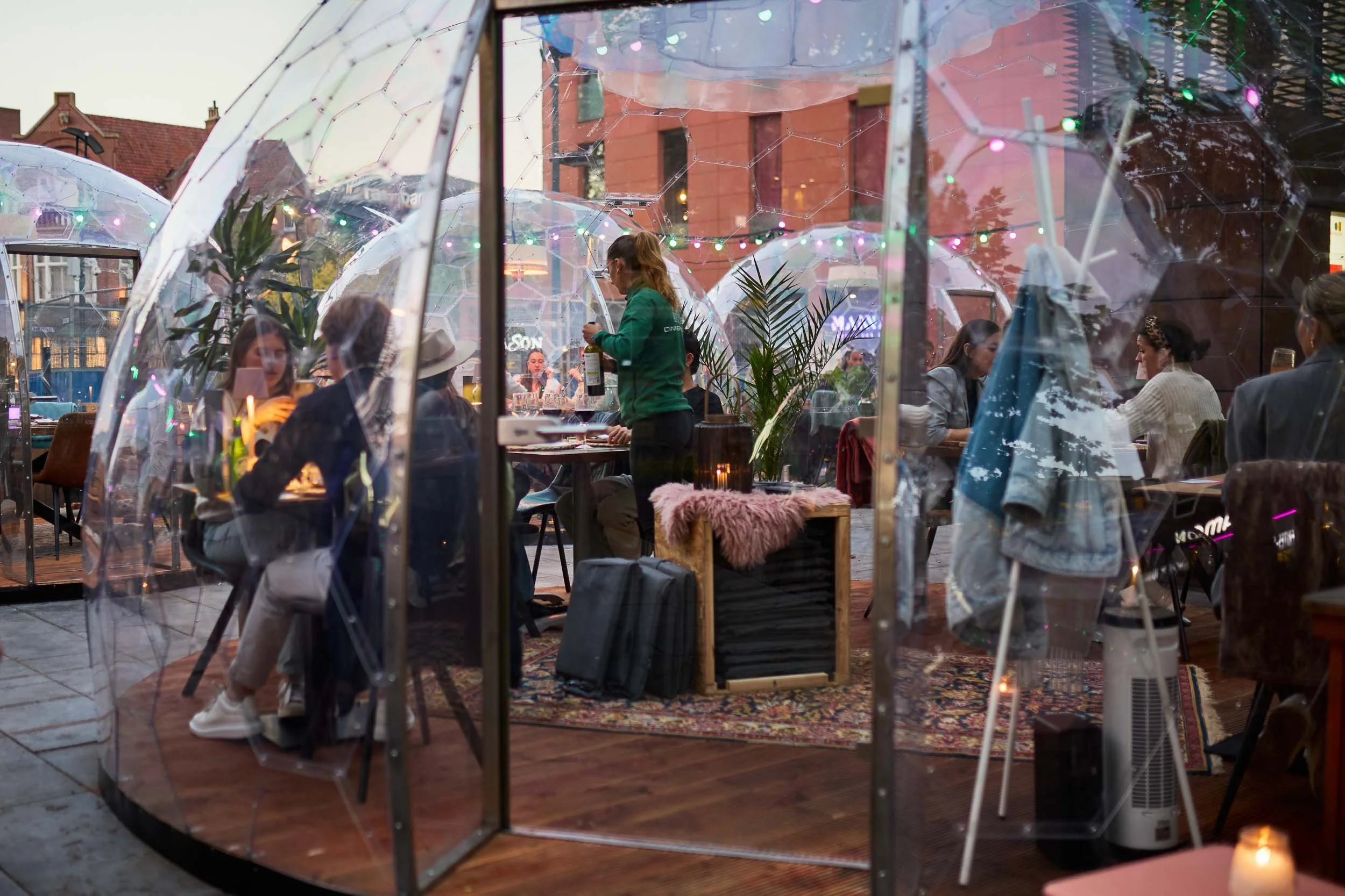 Binnen bij een restaurant of café met mensen die dineren, gezien door een glazen tent met decoraties en planten, met mensen die met elkaar praten en genieten van hun maaltijd.
