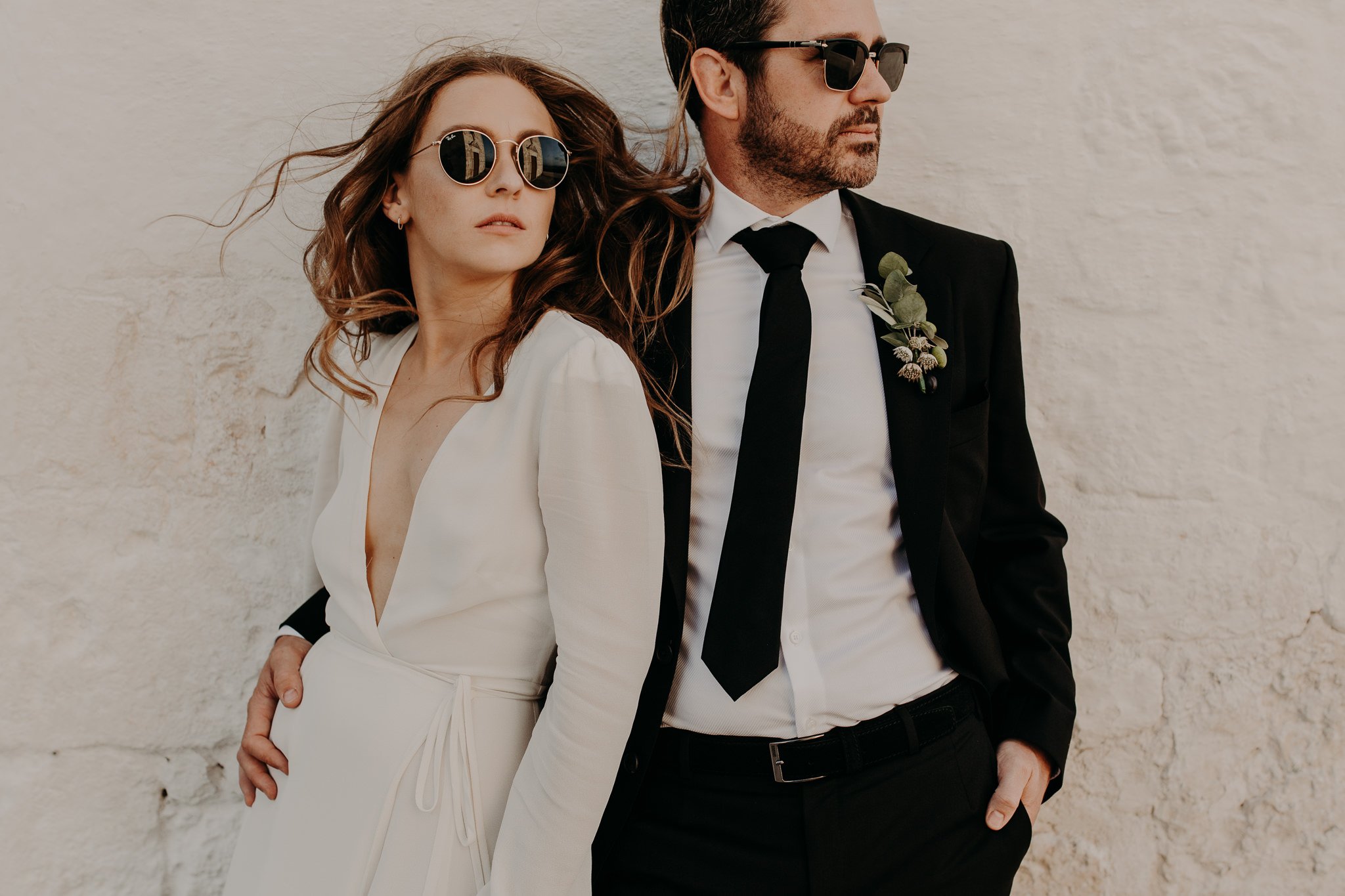 Un marié et une mariée posant devant un mur blanc et portant des lunettes de soleil