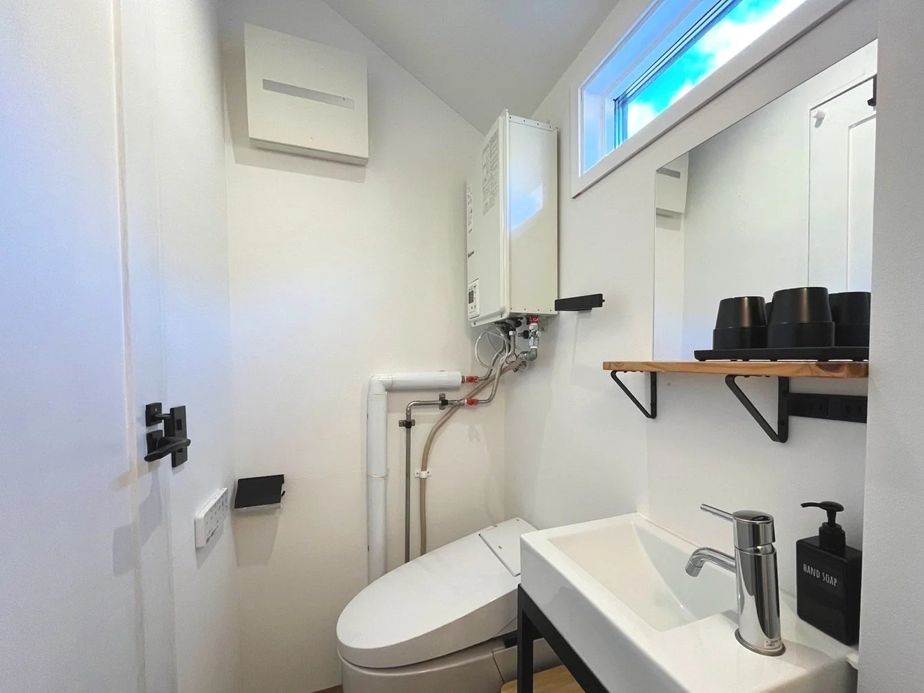 Small modern bathroom with a high-tech toilet, a small sink with a soap dispenser, a window with blue sky, and a wooden shelf with black cups.