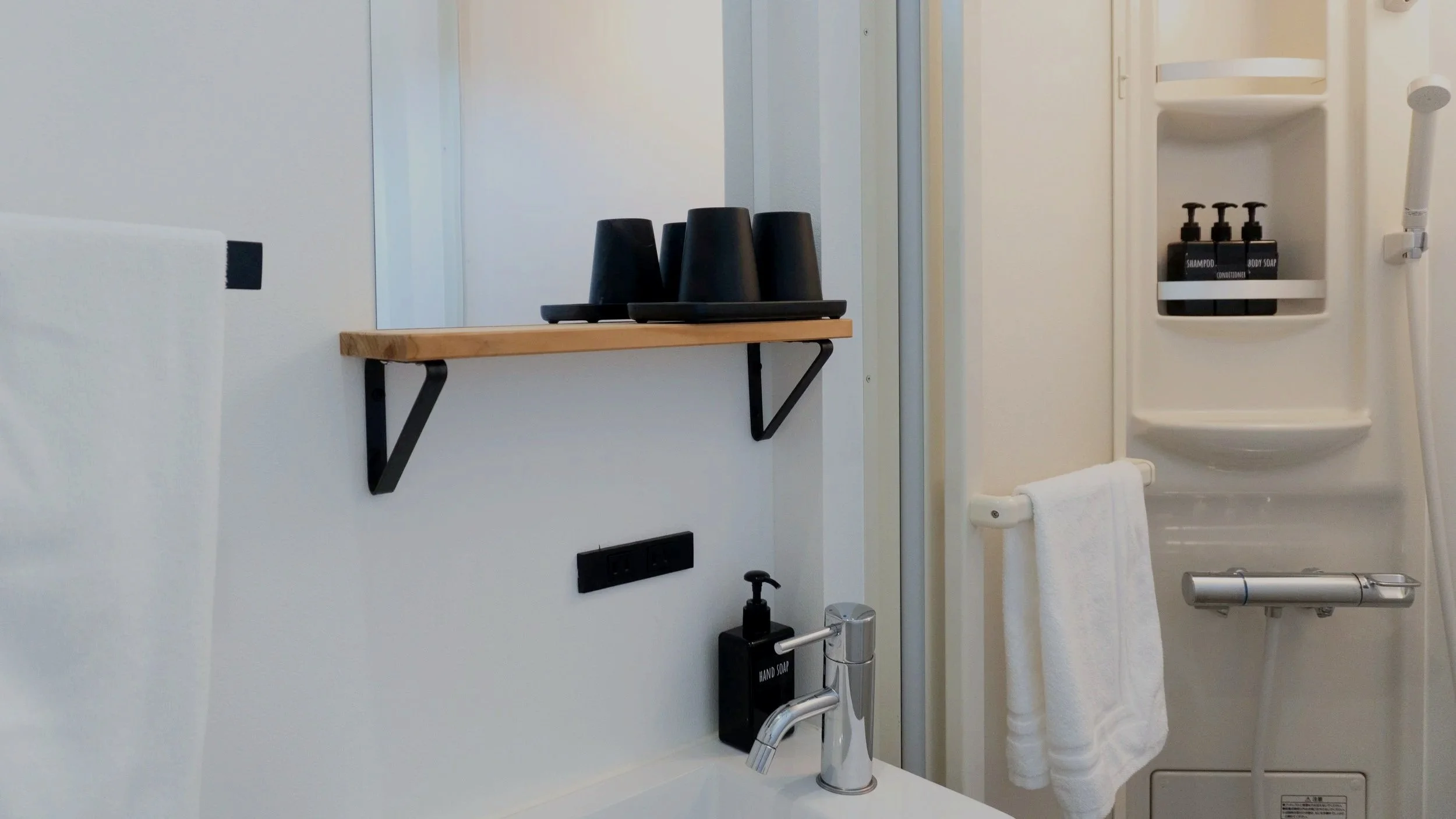 Bathroom with a mirror, black disposable cups on a wooden shelf, and toiletries including shampoo and body soap in a shower, a towel hanging on a rail, and a white washing machine with a showerhead.