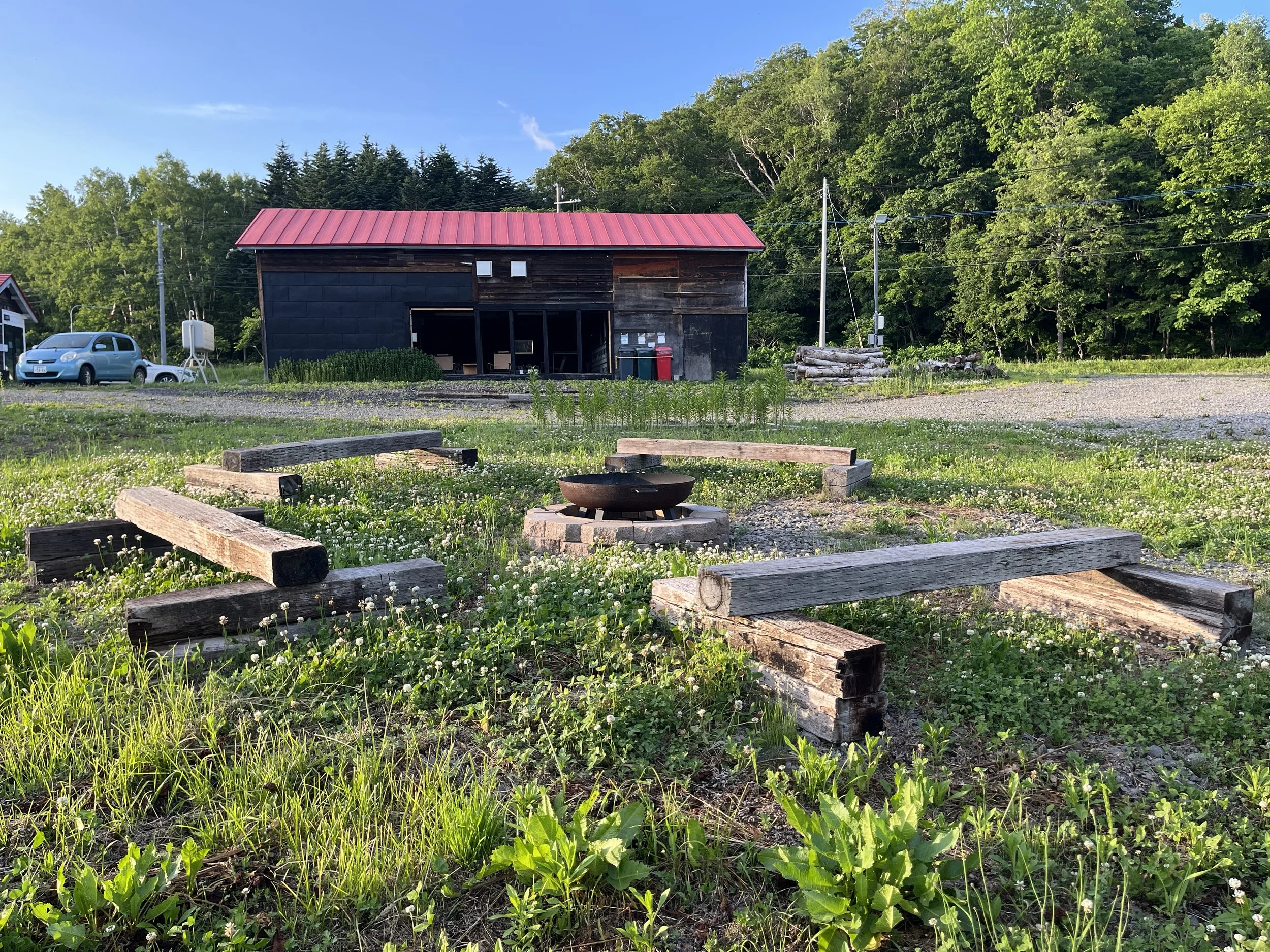 A rural scene with a BBQ house with a red metal roof surrounded by trees, parked cars, and a grassy area with a fire pit.