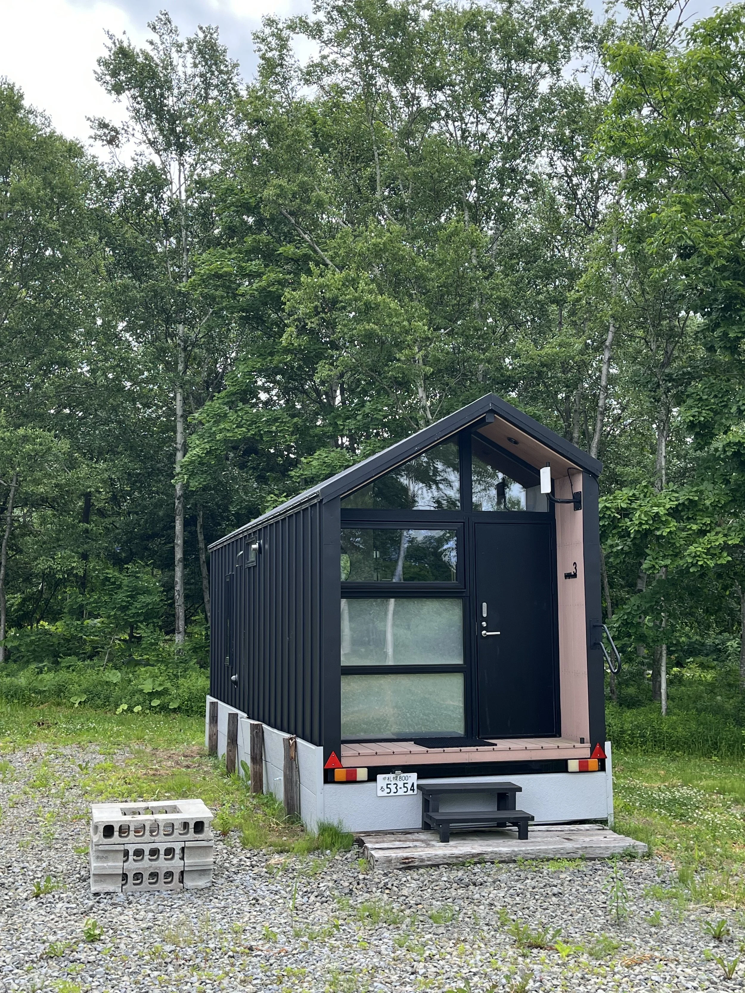 A black trailer parked on a gravel lot, with a background of green trees, and a cloudy sky.