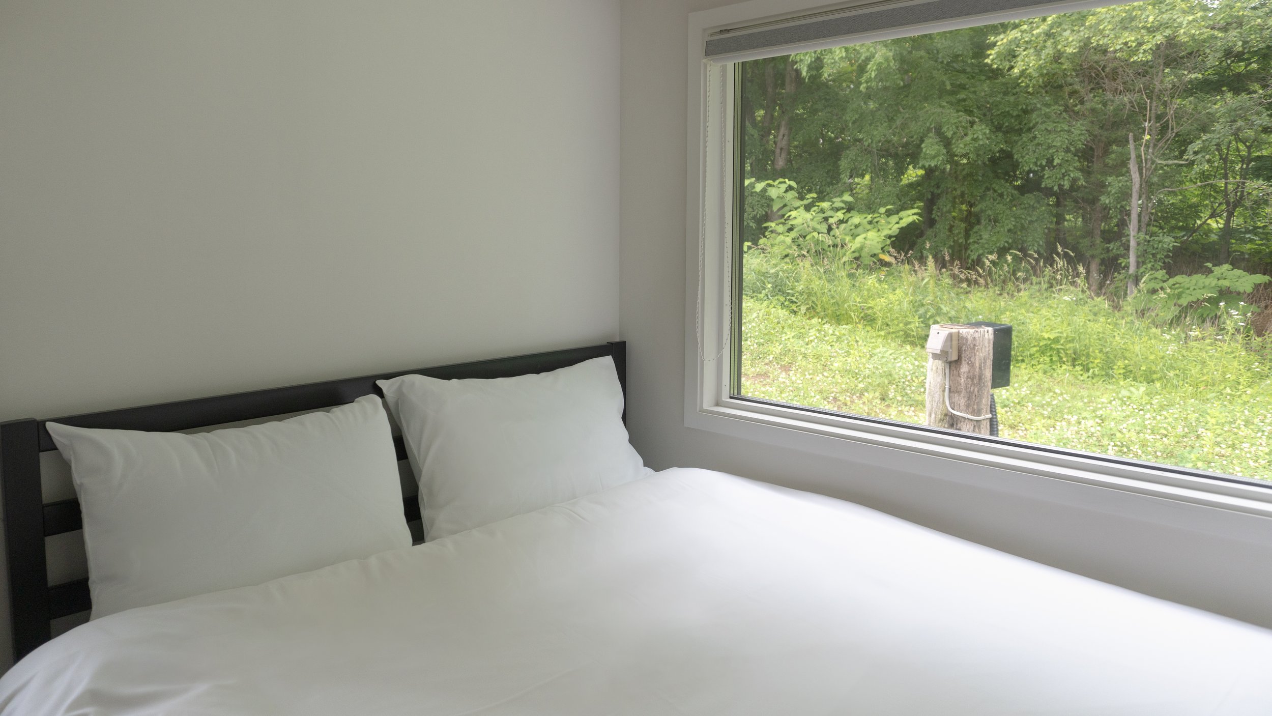 A bedroom with a bed featuring white sheets and pillows, next to a large window showing a green, leafy outdoor scene.