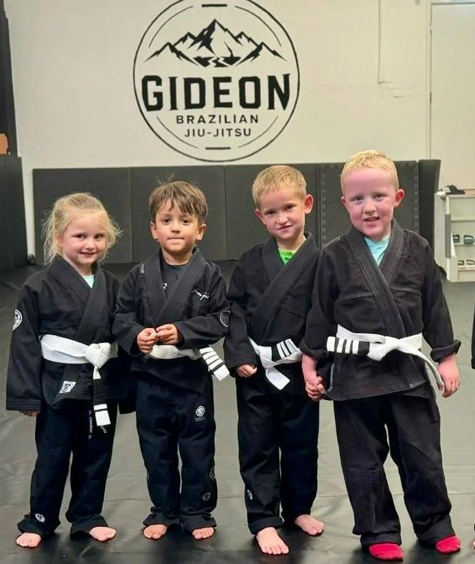 Four young children in black Brazilian Jiu-Jitsu uniforms with white belts standing together on a mat inside a gym, with a 'Gideon Brazilian Jiu-Jitsu' logo on the wall behind them.