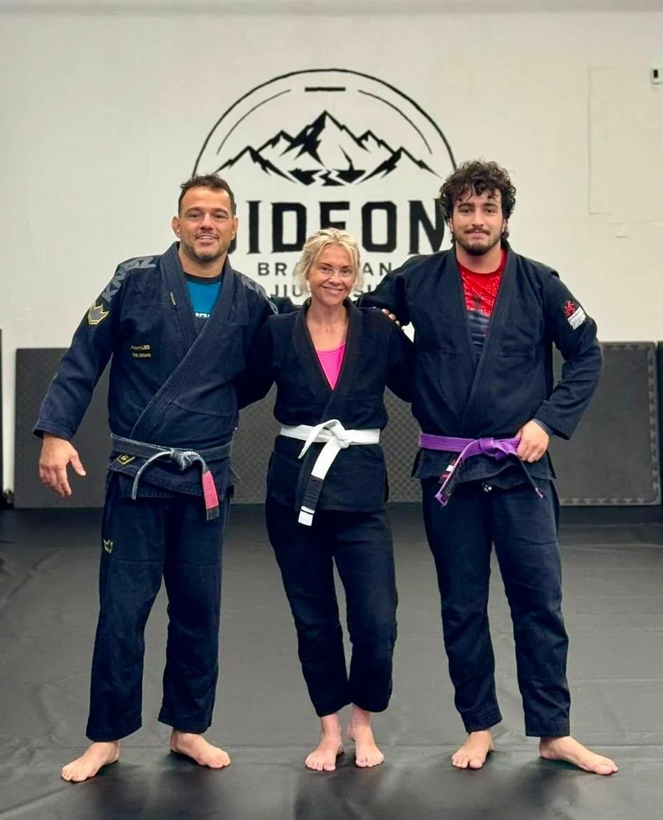 Three people wearing Brazilian Jiu-Jitsu gis standing in a gym with a logo of a mountain and the words "IDF Jiu Jitsu" in the background. The person in the middle is a woman with blonde hair and a white belt. The two men on either side are men with dark hair and colored belts, one blue and one purple.