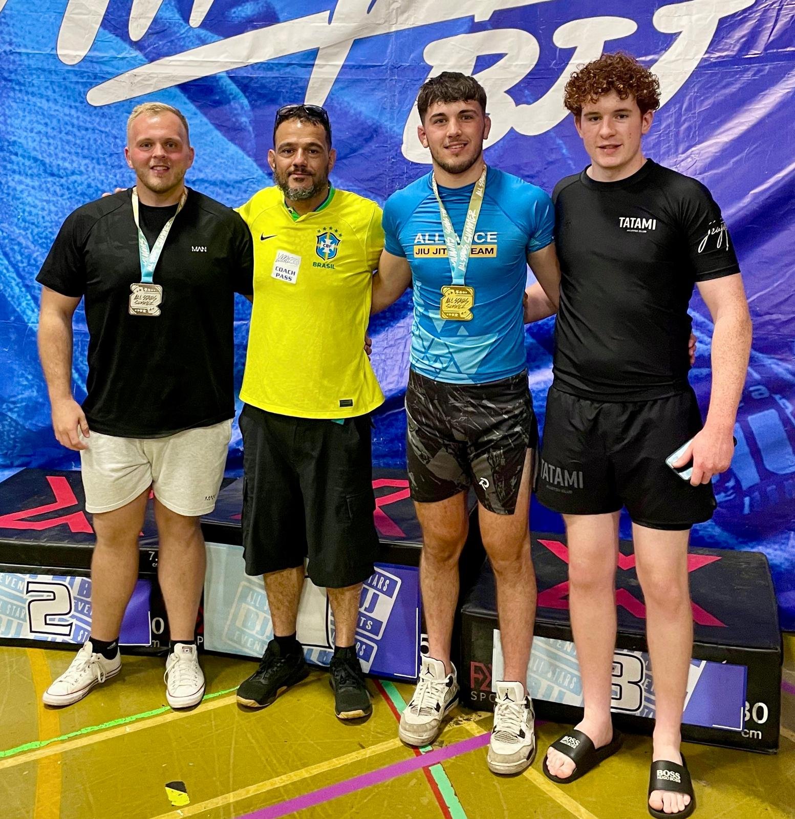 Four men standing on a winners' podium with medals around their necks, smiling at the camera. The second man from the left is wearing a yellow Brazil sports jersey, the third man is wearing a blue athletic shirt, and the fourth man is wearing a black Nike shirt. The background features a blue banner and a sports event setting.