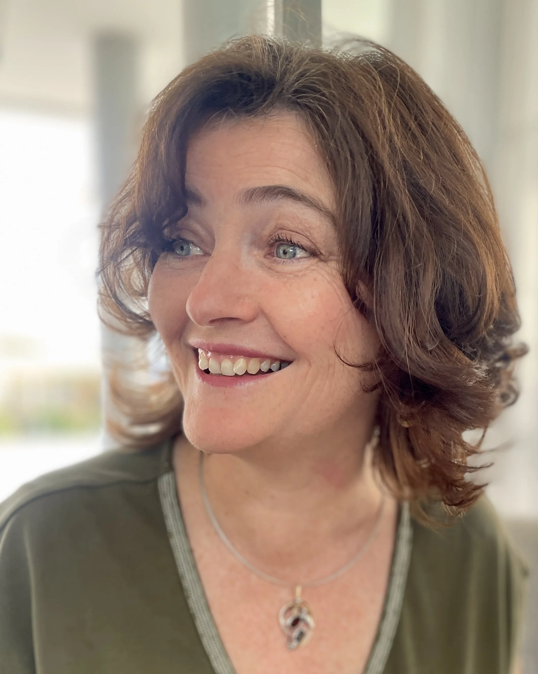 Close-up of a woman with short, wavy brown hair smiling and looking to her left, wearing a green top and a silver necklace.