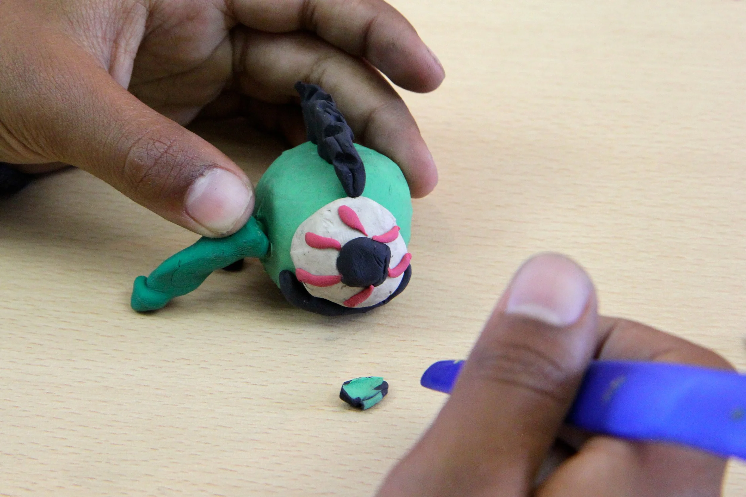 Hands making a small clay model of a person or creature on a wooden table.