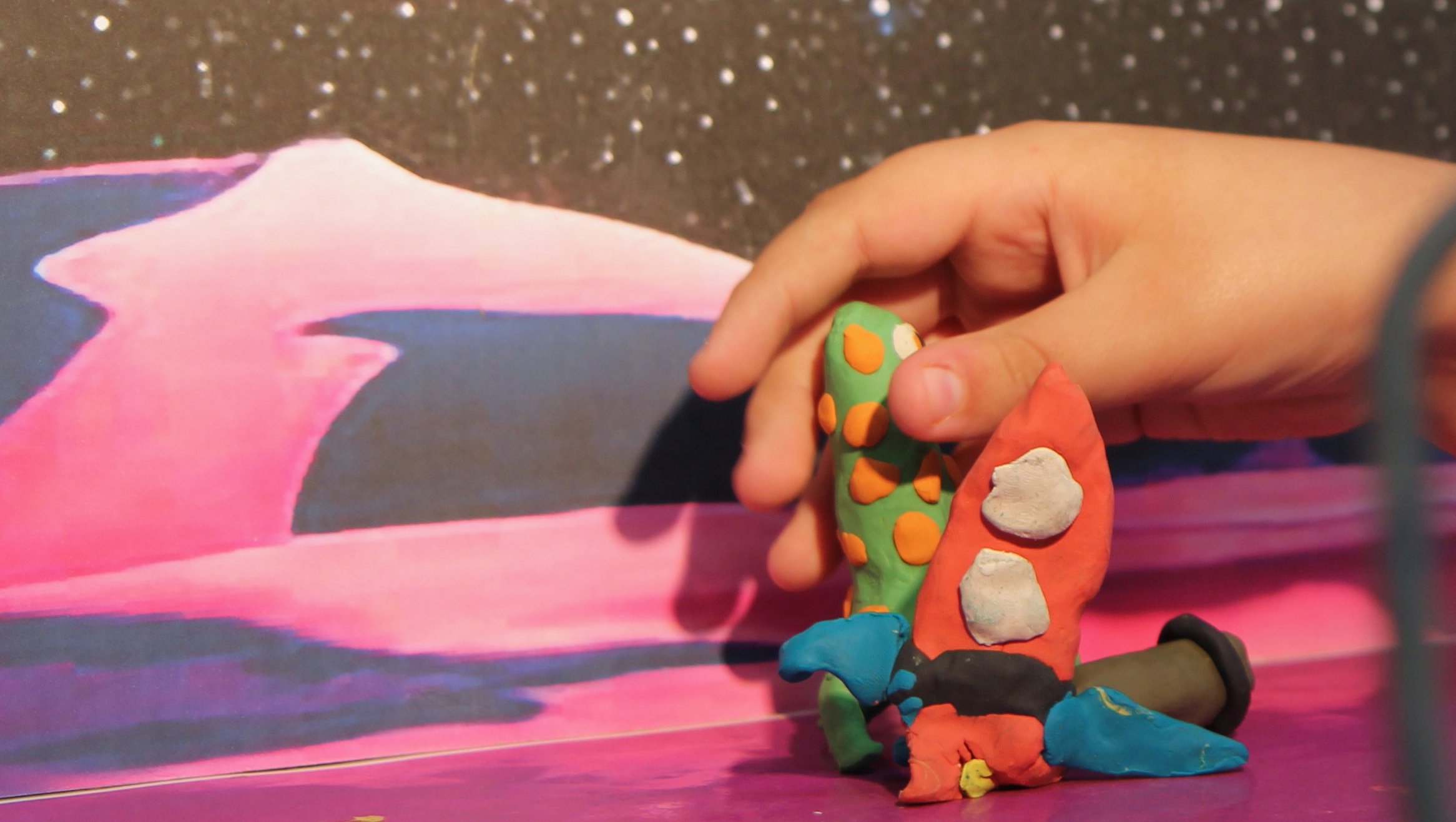 Hand holding colorful clay figures with a painted pink mountain and starry sky background.