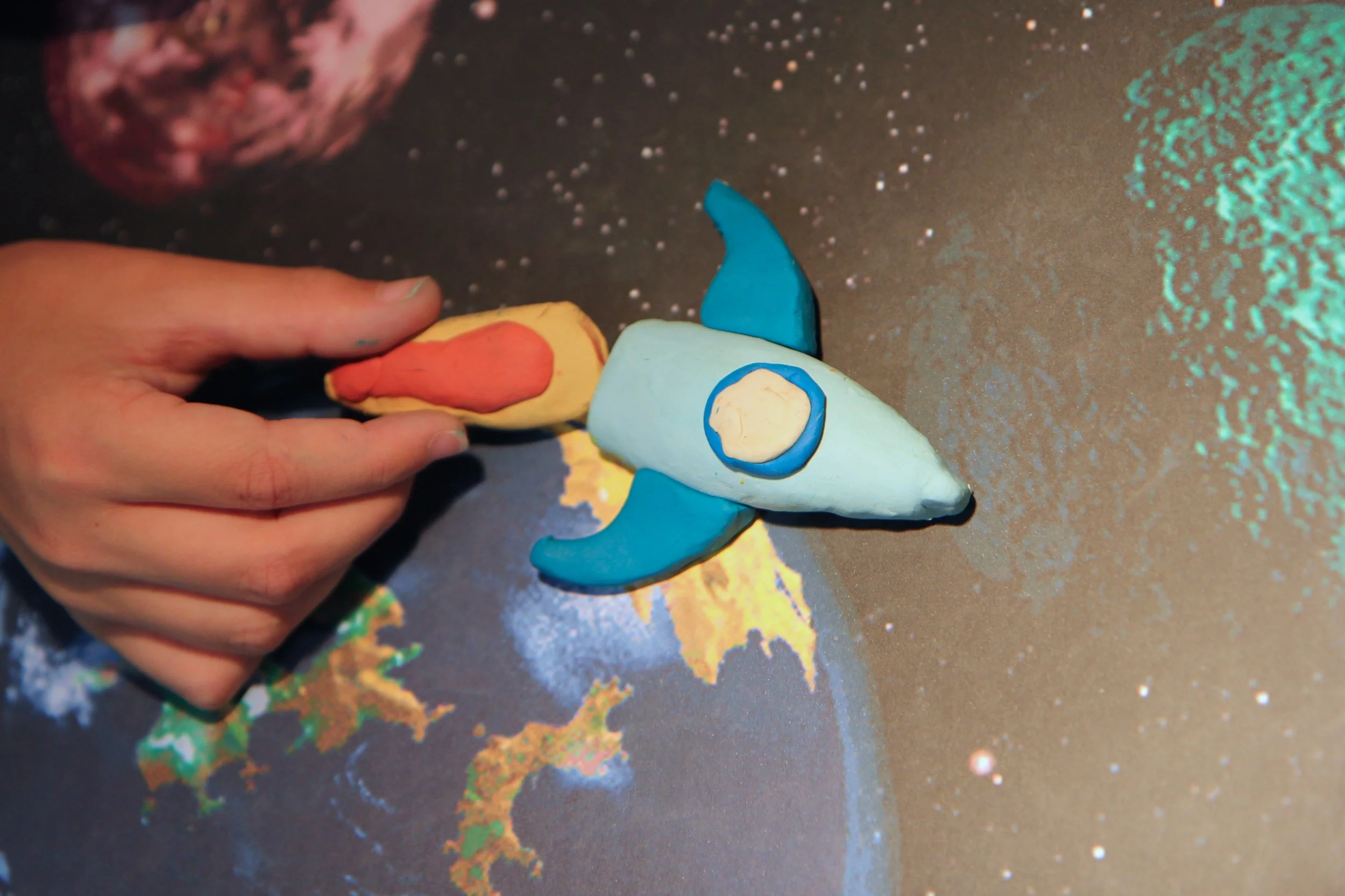 A person holding a colorful clay model of a rocket with blue fins, a white body, and red-orange accents, placed on a space-themed surface with planets and stars in the background.