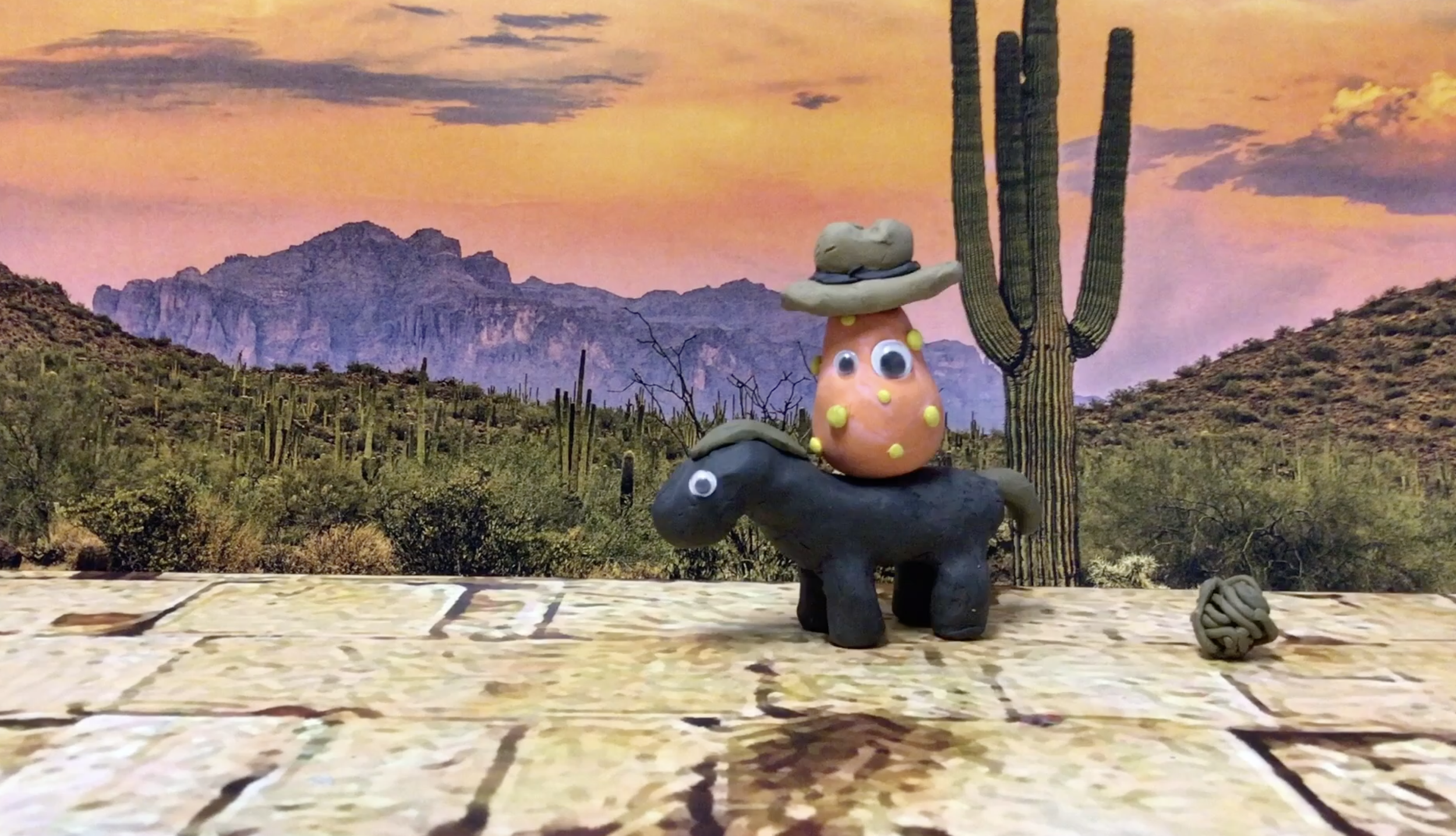 A clay sculpture of a creature with googly eyes and yellow dots sits on a black horse made of clay, with a cactus and a sunset mountain background.