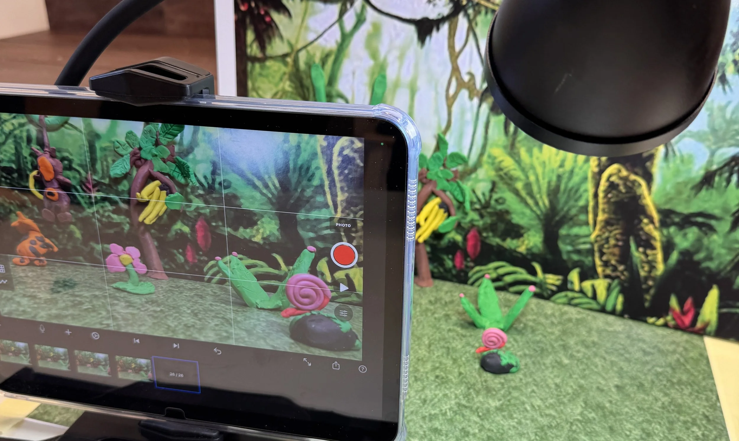 A tablet is recording a scene of colorful clay models of snails, ladybugs, and plants in a diorama background that resembles a forest scene.