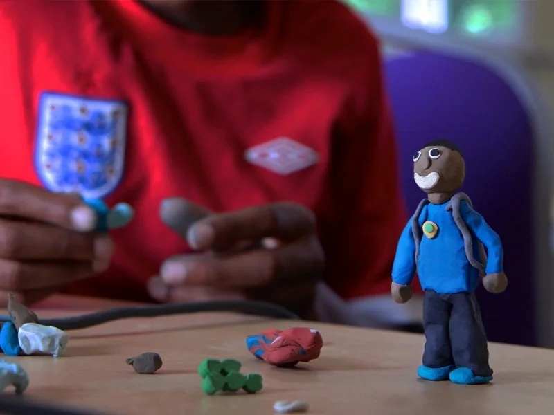 Close-up of a person in a red shirt with a blue and white badge, working on a blue mascot figurine with a grey backpack and glasses, in a setting with colorful lights. Several small clay sculptures are on the table.