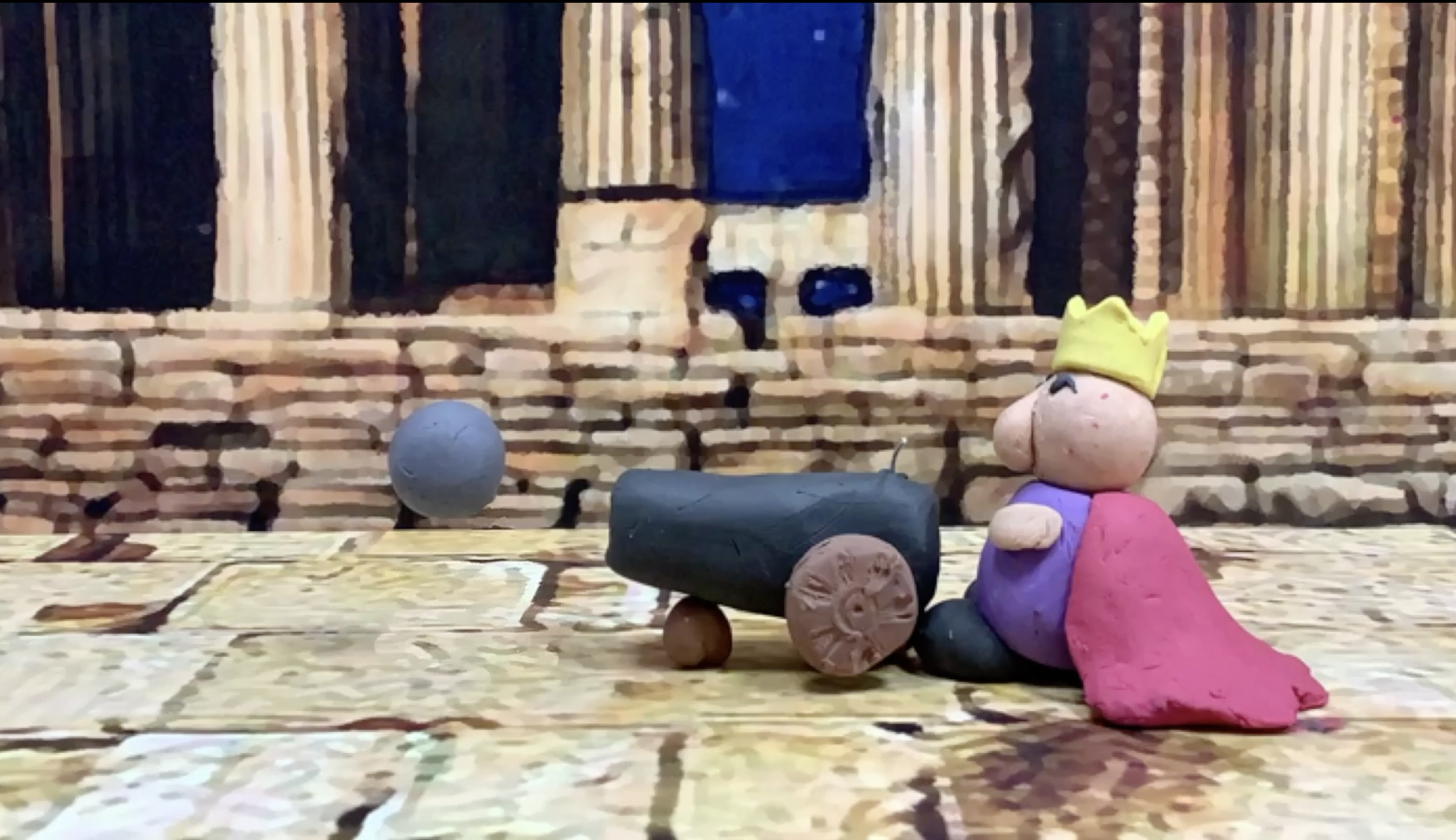 A small clay figure of a king with a yellow crown, sitting beside a black cannon, with a blue sphere nearby, on a wooden surface in front of a brick background.