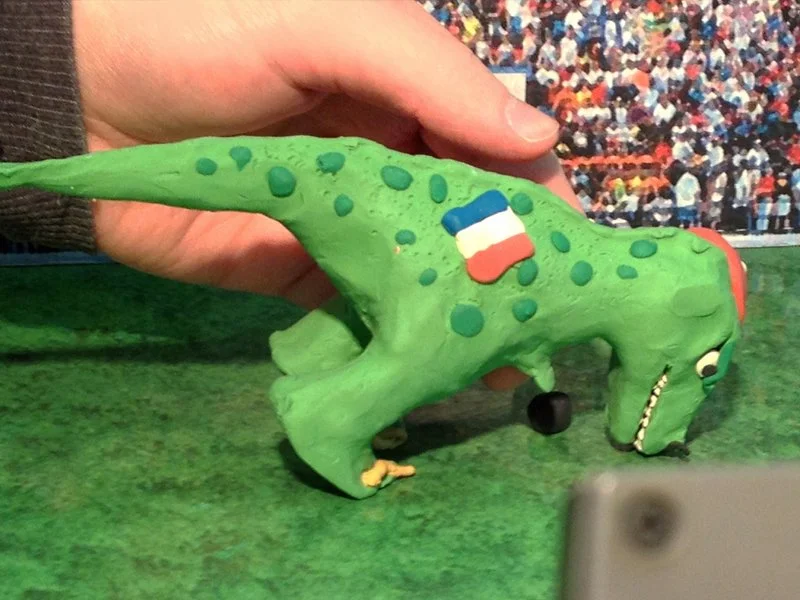 A small green model of a dinosaur with a long tail, patterned with blue spots, and a face featuring a large eye with an eyelid. There is a cartoonish French flag on its side. The dinosaur is placed on a green surface with a colorful crowd in the back