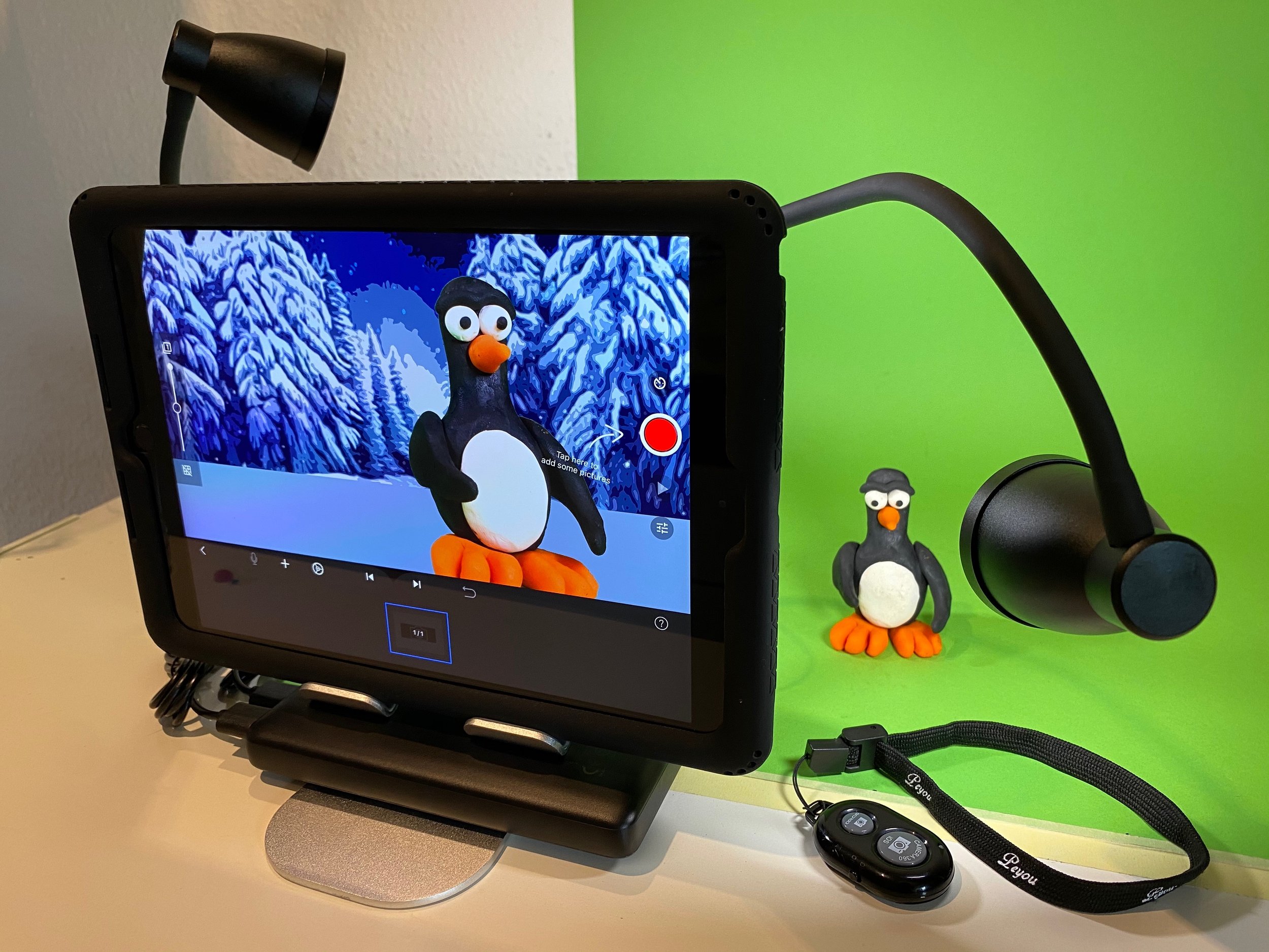 A tablet on a stand displaying a cartoon penguin with an orange beak and feet, in front of a snowy, wintery background. To the right of the tablet, there is a small figurine of the same penguin and a black wireless microphone with a headband and remo