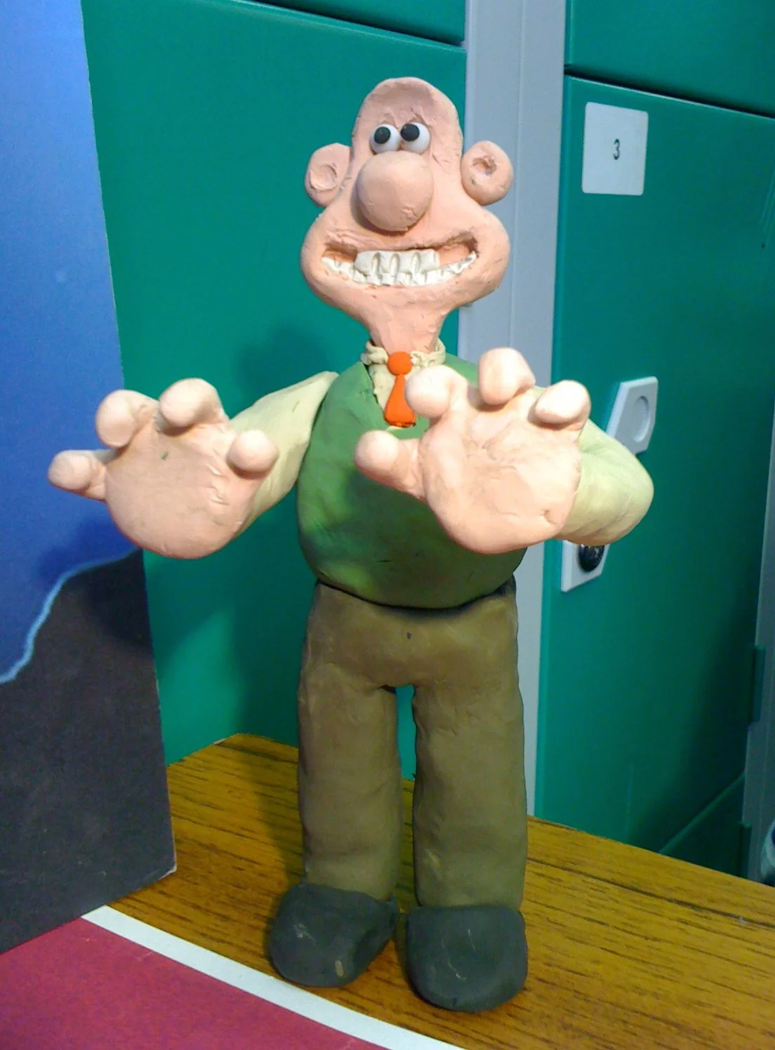 Clay sculpture of a cartoon character with big eyes, large nose, wide teeth, wearing a green shirt, brown pants, and black shoes, standing on a wooden surface.