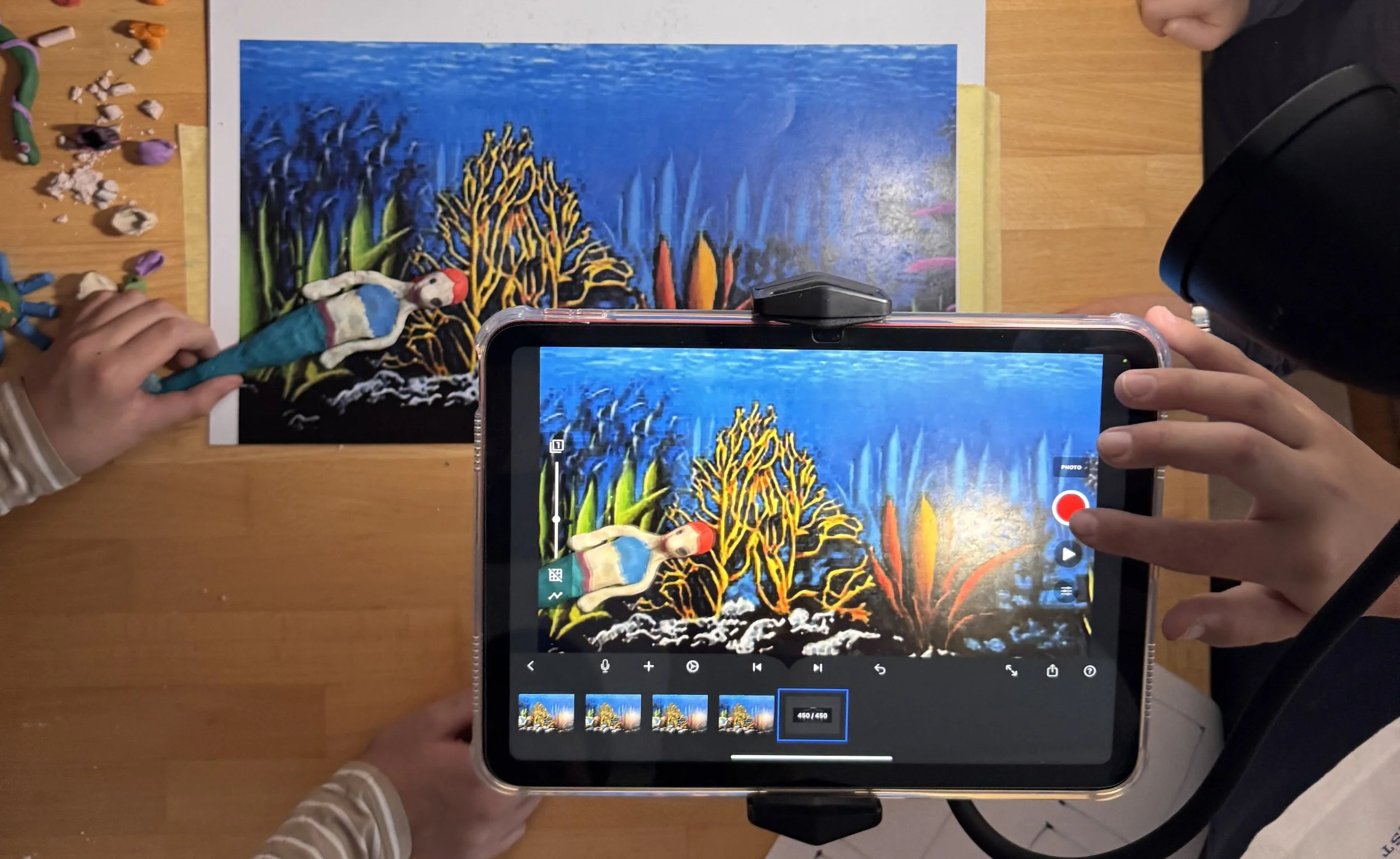 Children creating a mixed media art piece of a coral reef scene on paper, with one child holding a fabric puppet near the artwork and another taking a photo with a tablet.