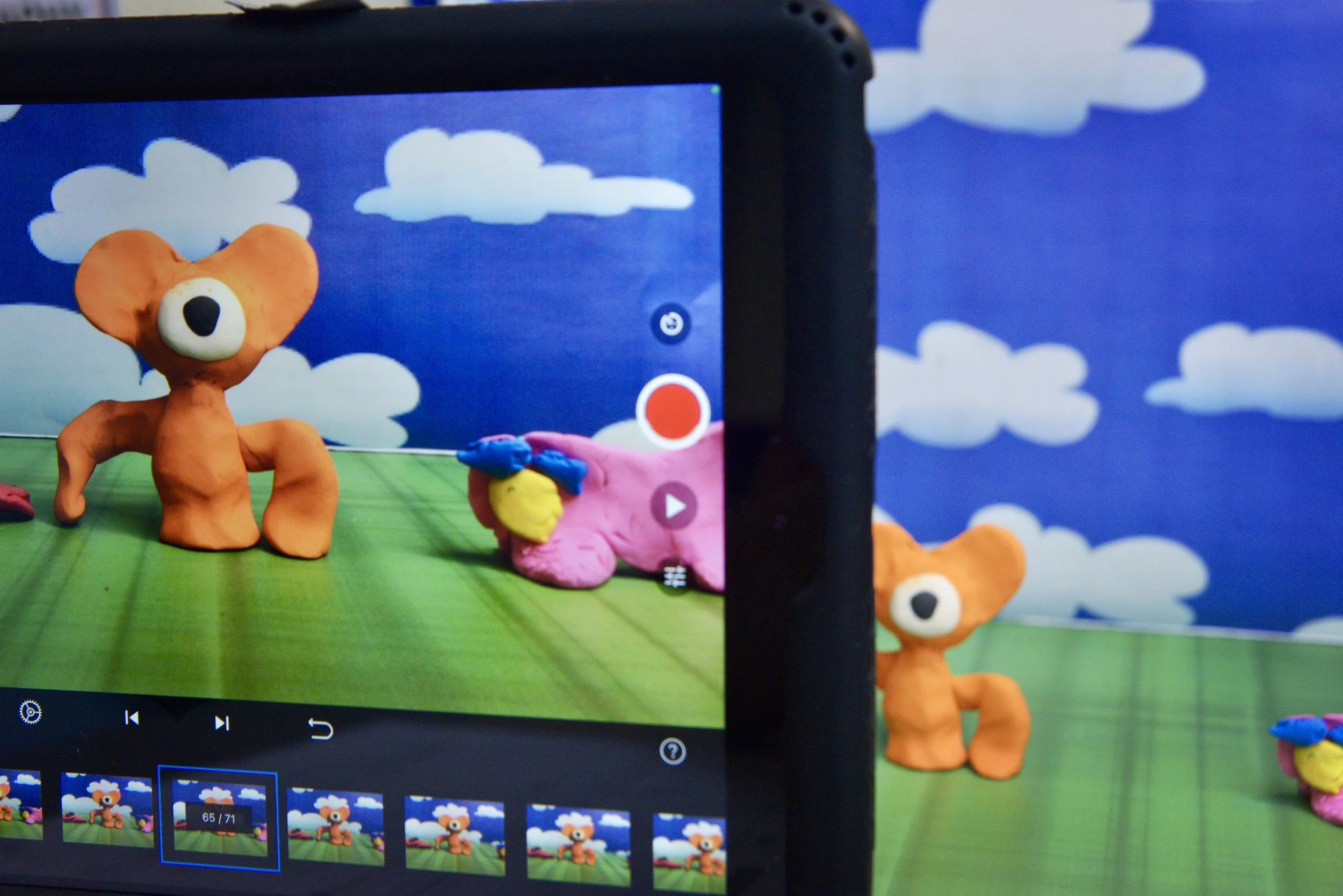 A scene with colorful clay figures and clouds in the background, seen through a smartphone screen recording a video of the figures.