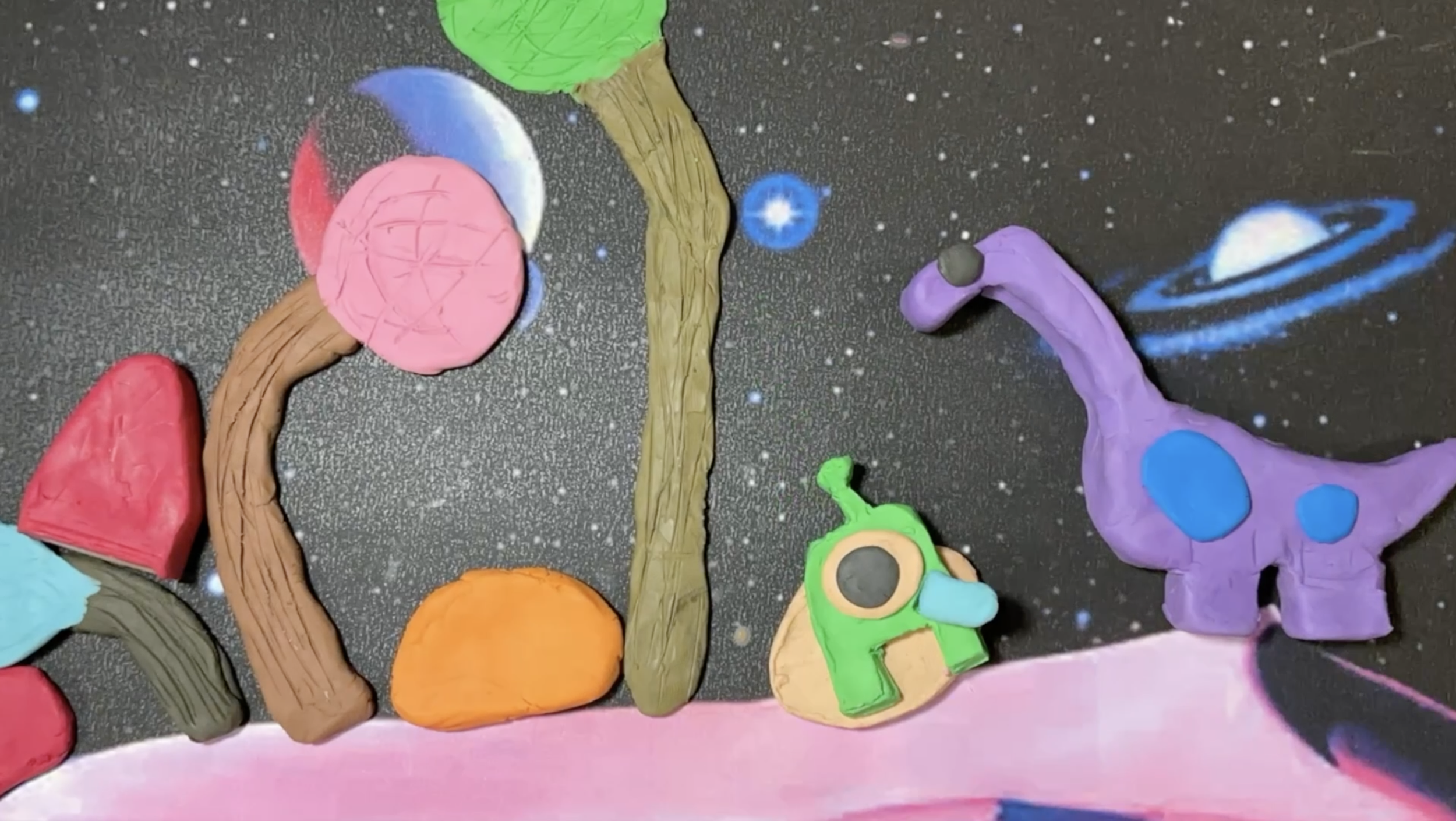 Colorful clay models arranged to spell the word 'LOVE' with a space-themed background featuring planets, stars, a galaxy, and a pink ground.