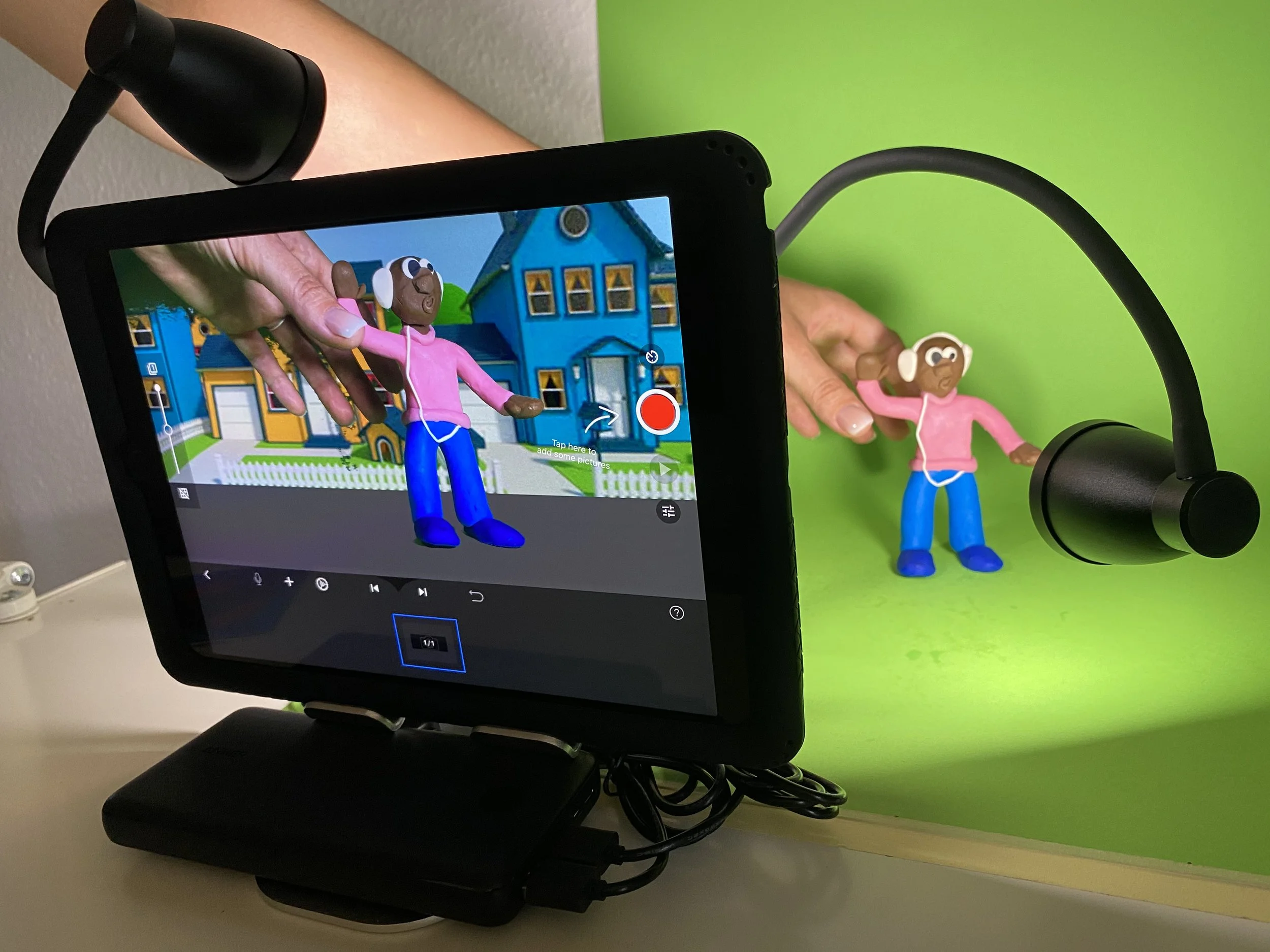 A tablet with a camera recording a stop-motion animation of a clay figure of a girl with brown skin, wearing a pink hoodie and blue pants, on a green background. The clay figure is also held in front of the tablet for filming.