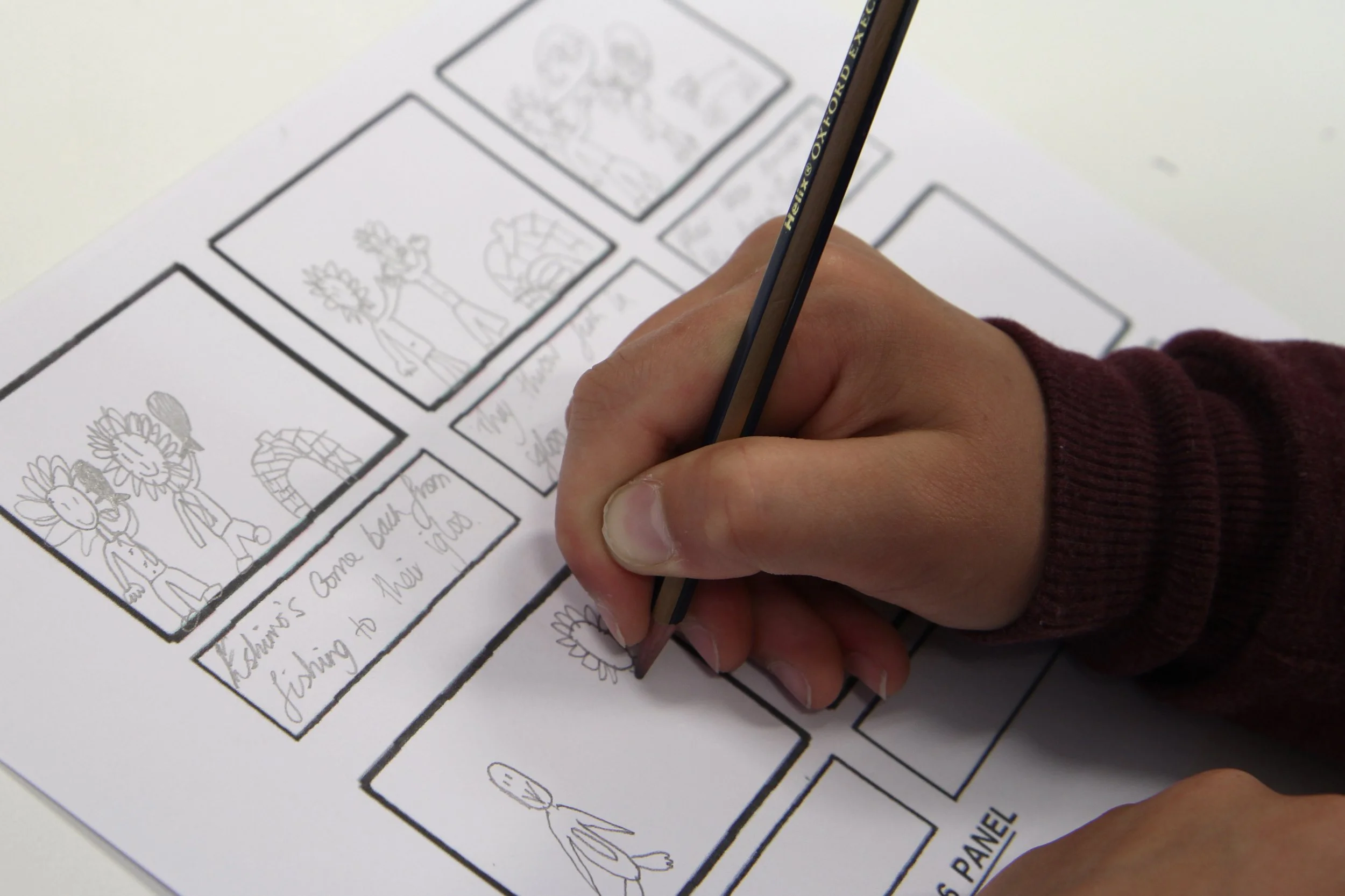 Child's hand coloring a sunflower in a comic strip storyboard with black ink outlines, with sketches and handwritten notes on the paper.