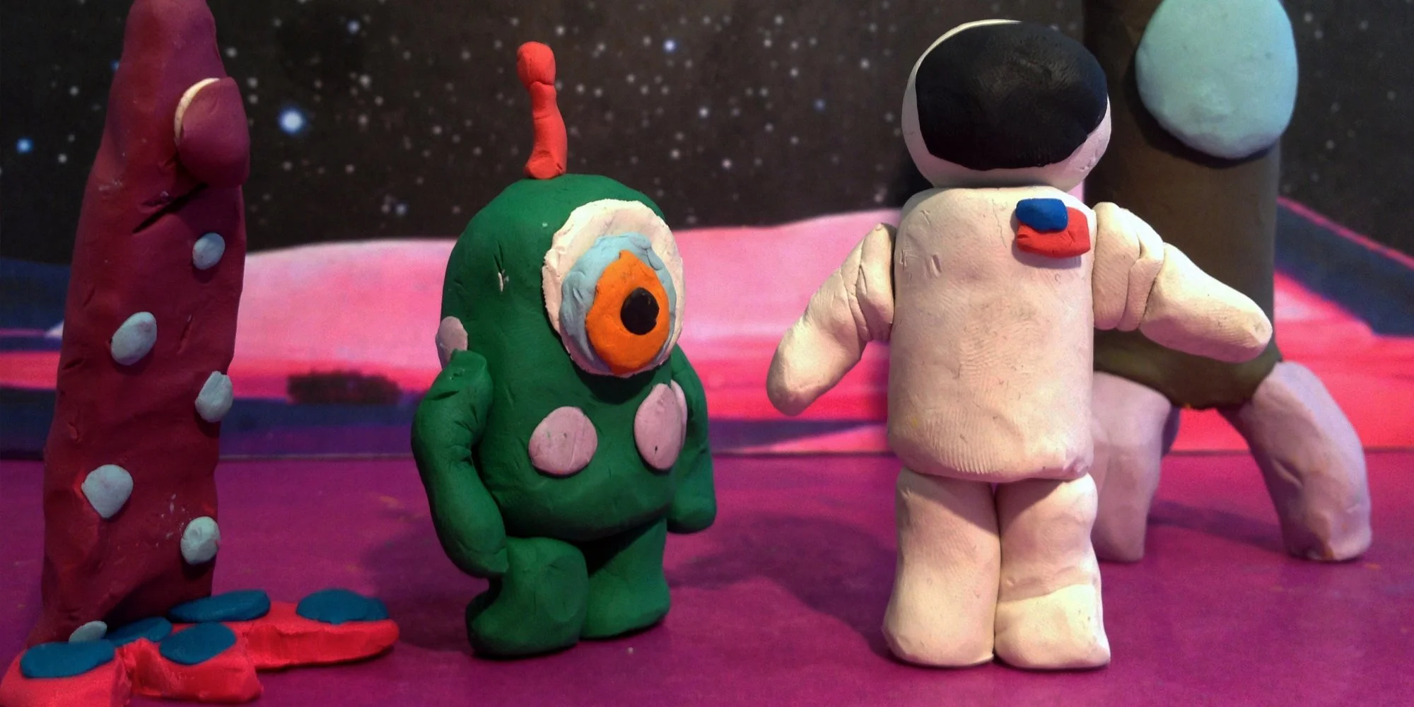 Colorful clay models of an alien, a human astronaut, and a red and blue alien are displayed against a space background with a planet and stars.