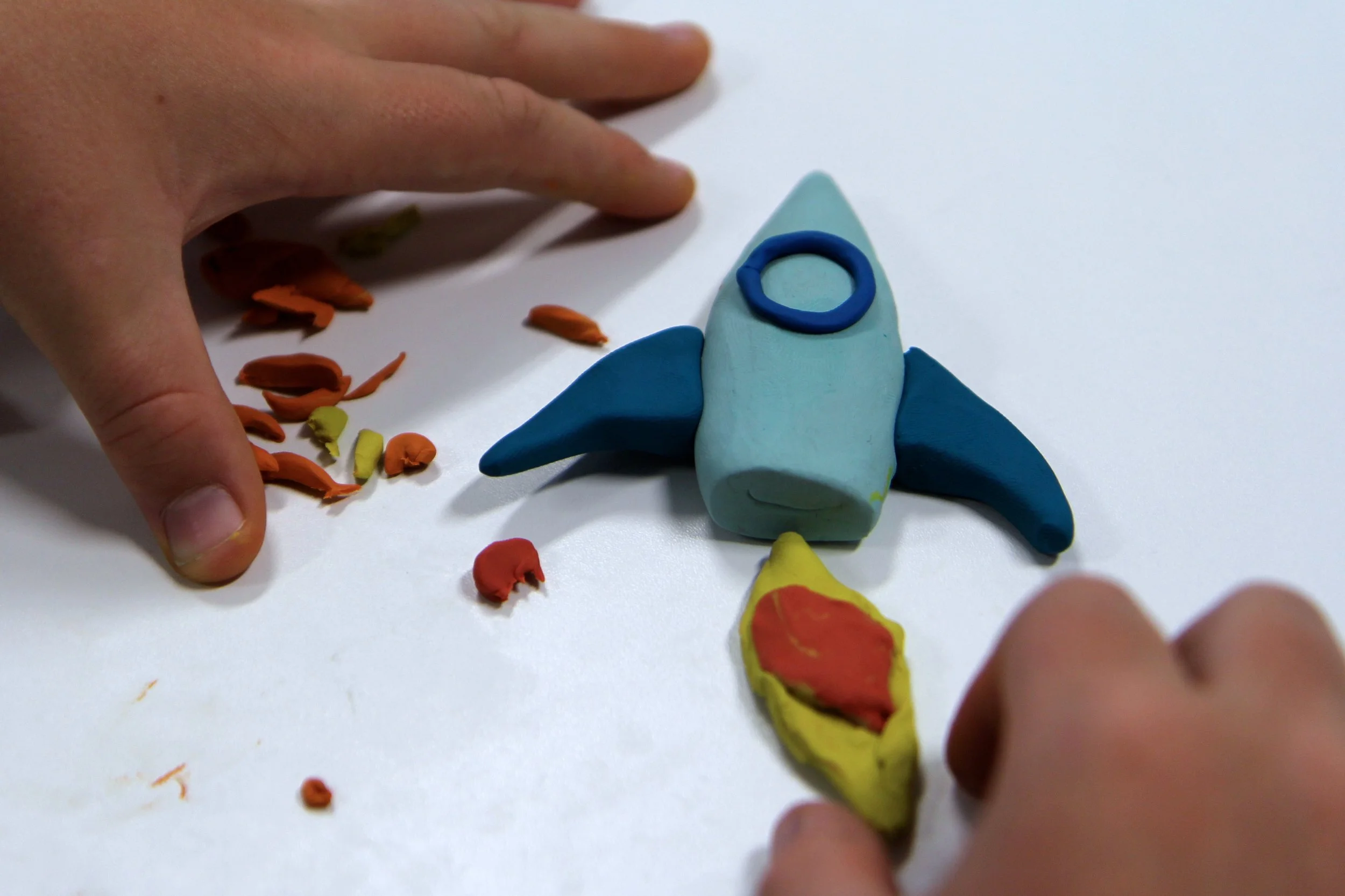 A child's hands working on a colorful clay model of a rocket ship with a red and yellow flame, surrounded by scattered clay pieces on a white surface.