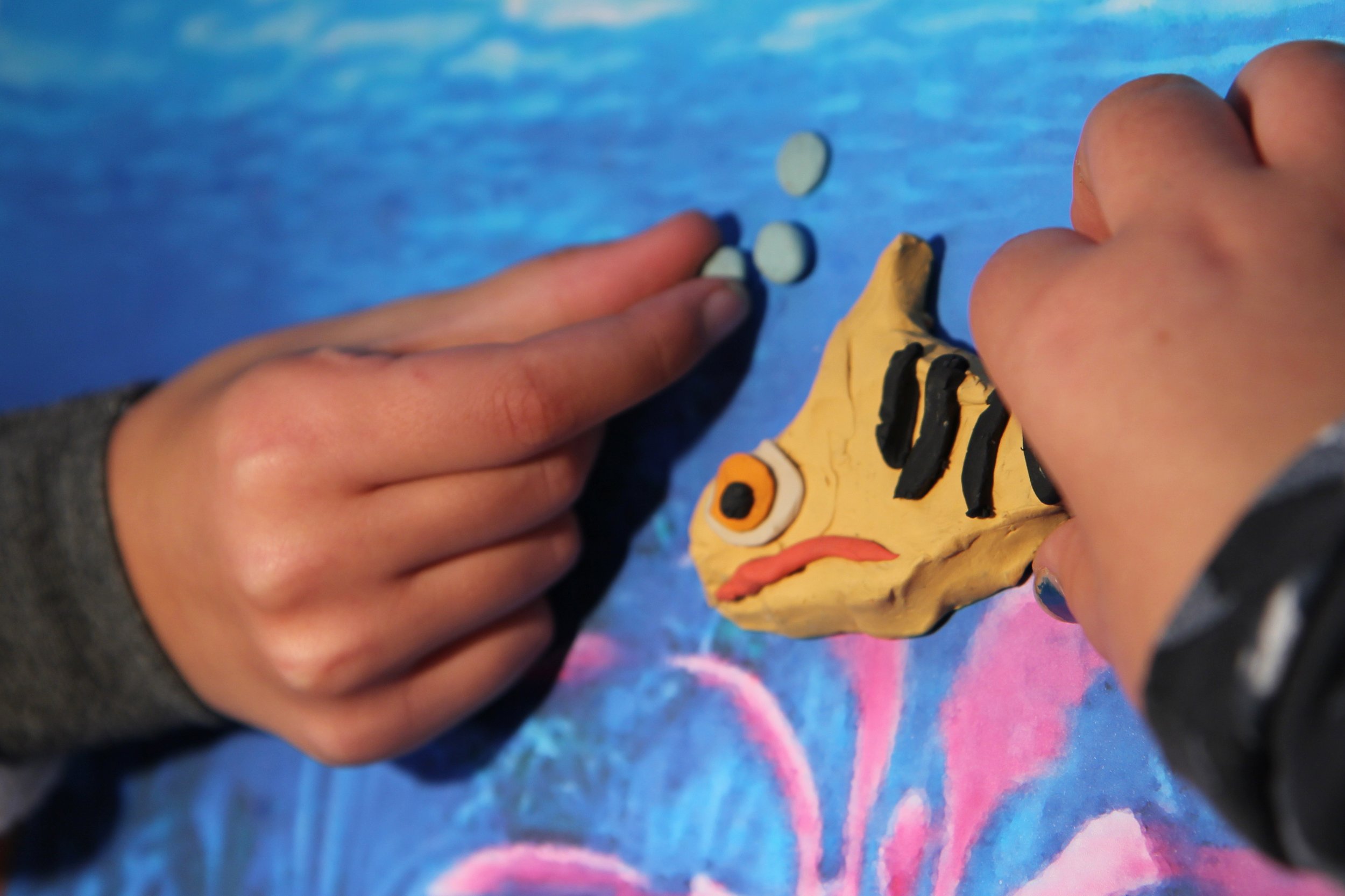 Child's hands playing with a clay fish and small round objects on a colorful surface.