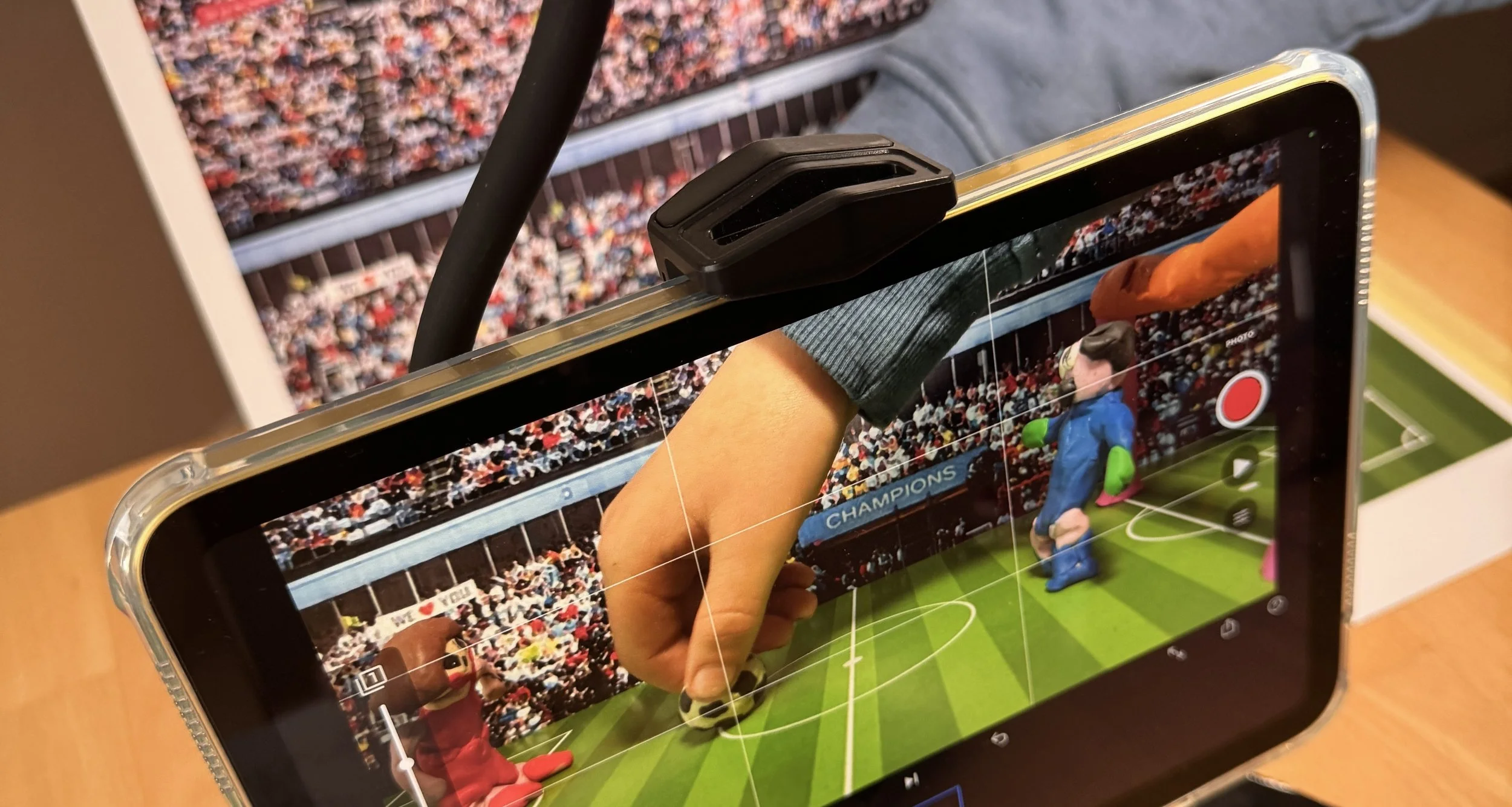 A smartphone showing a video recording of a man’s hand, a toy mascot, and a soccer field with a crowd in the background.