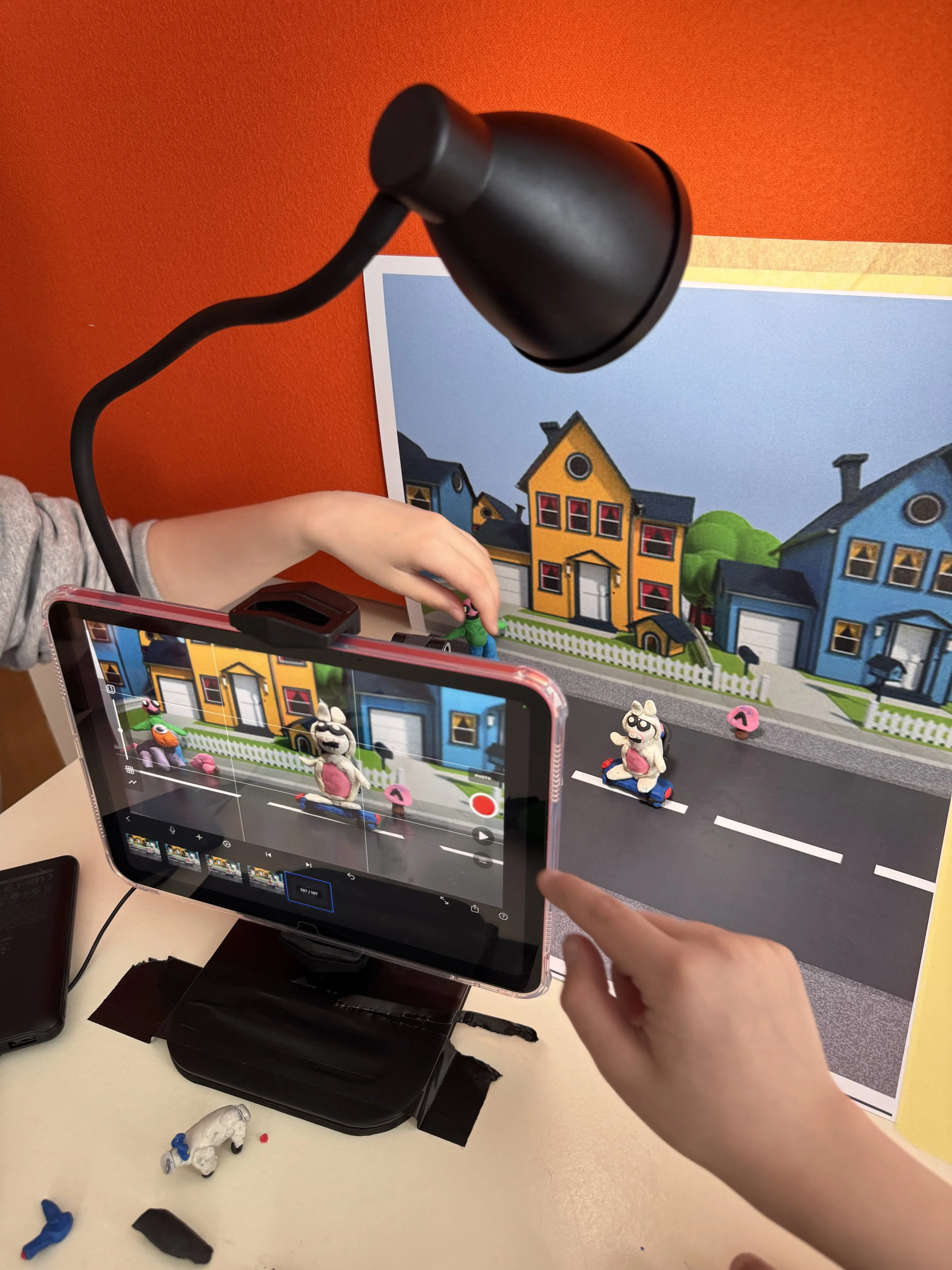Children filming a puppet show with toy puppets on a miniature road set, with a colorful house backdrop and professional lighting.