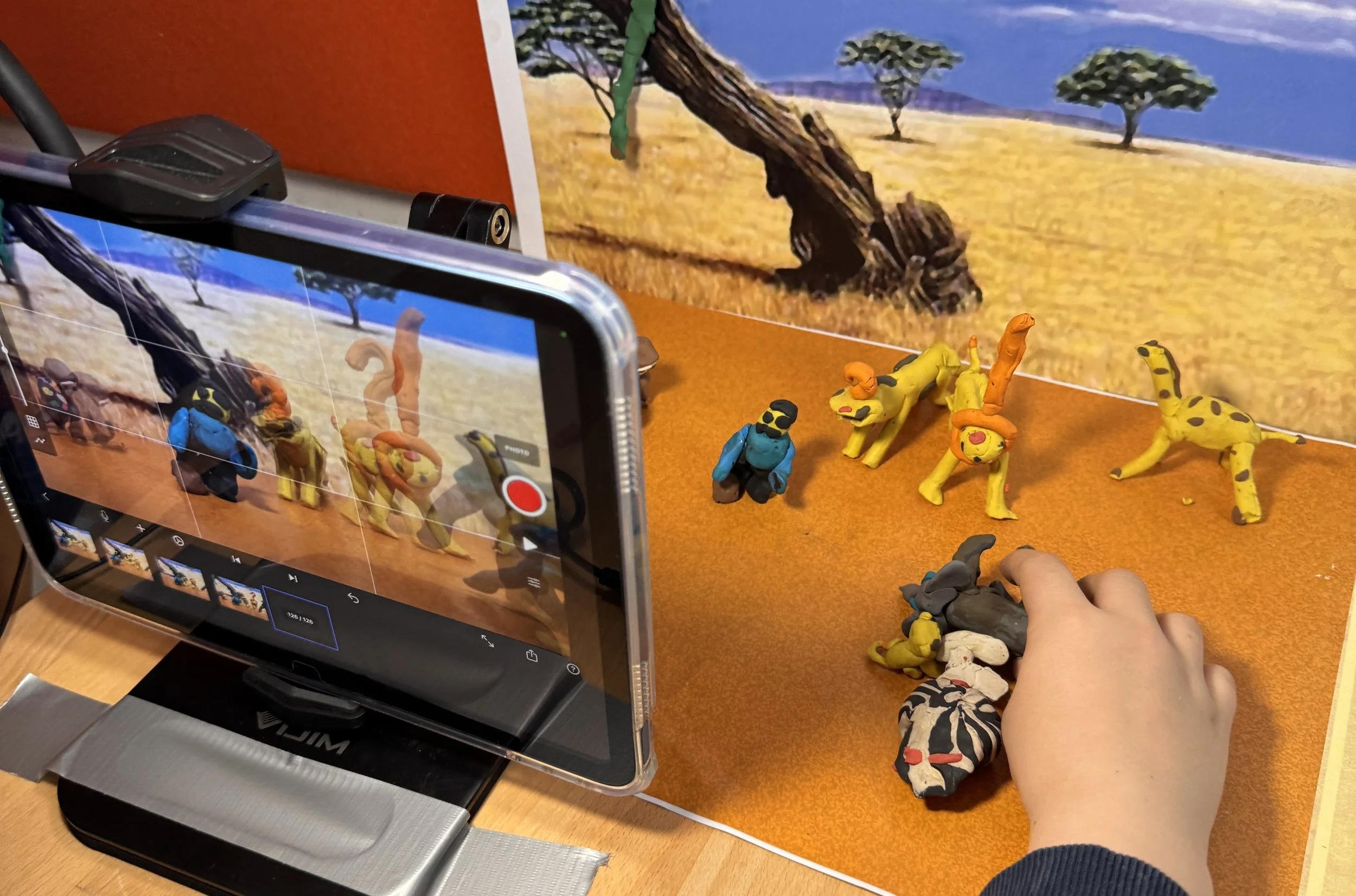 Close-up of a smartphone recording a scene of clay animal figurines, with a backdrop of a painted safari landscape featuring trees and a savannah. The clay animals include lions, a giraffe, and other wildcats, and a person's hand is adjusting or movi