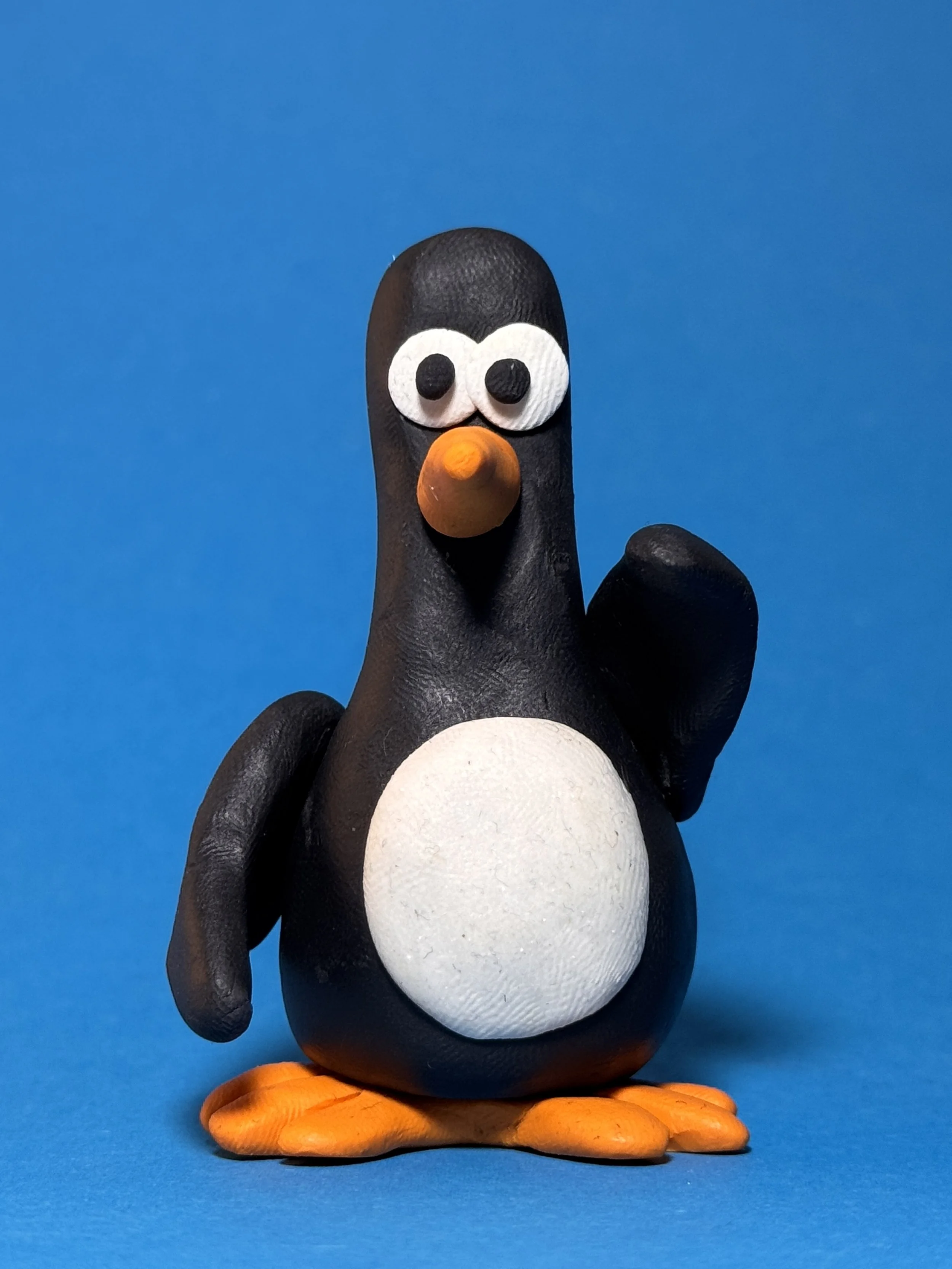 A small, cartoonish penguin figurine with a black body, white belly and face, orange beak, and orange feet, standing against a blue background.