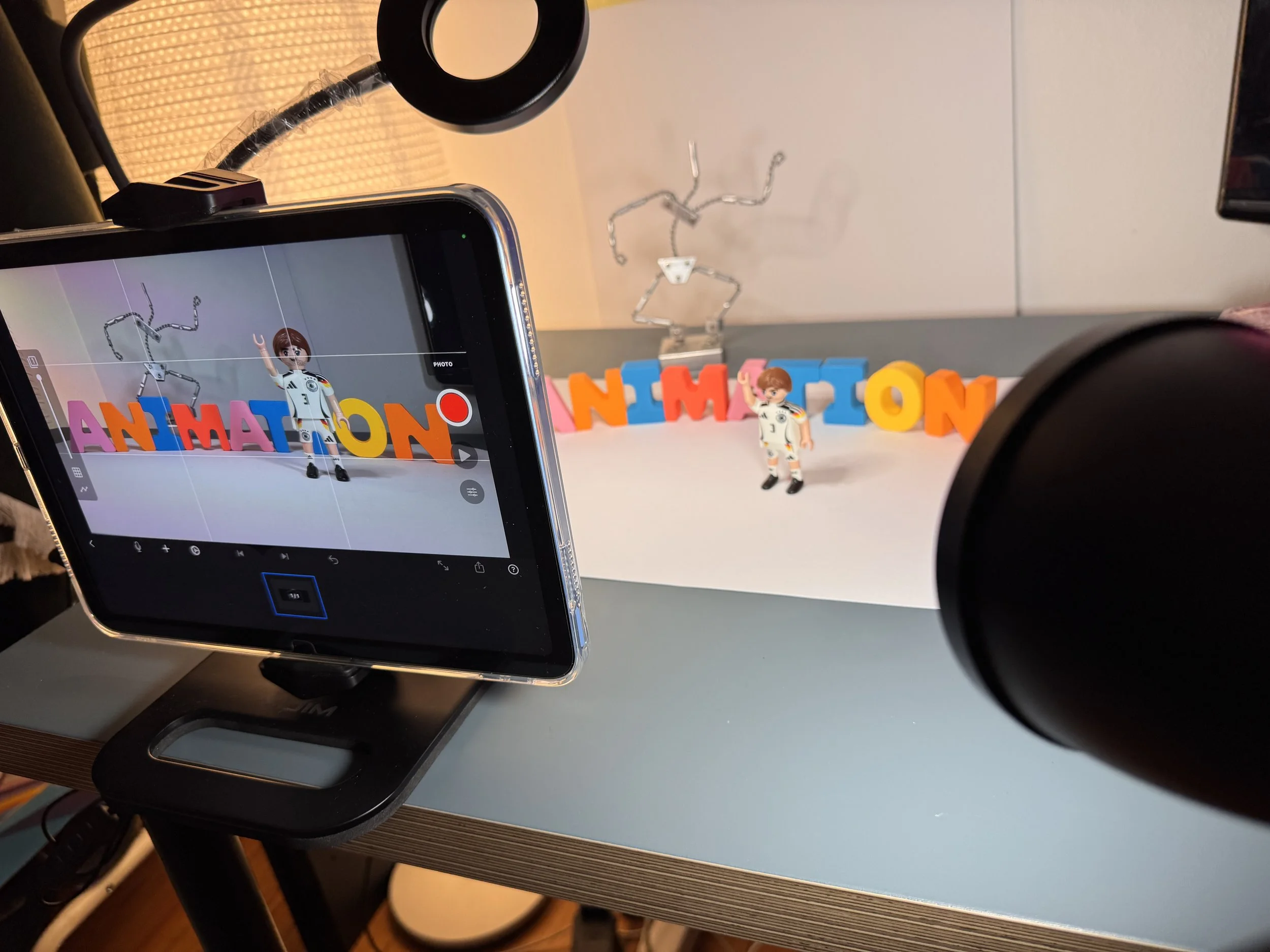 A smartphone mounted on a stand capturing a photo of two small toy figures in front of colorful letter blocks spelling 'ANIMATION' on a white surface. A ring light and a metallic wire sculpture are also visible in the background.