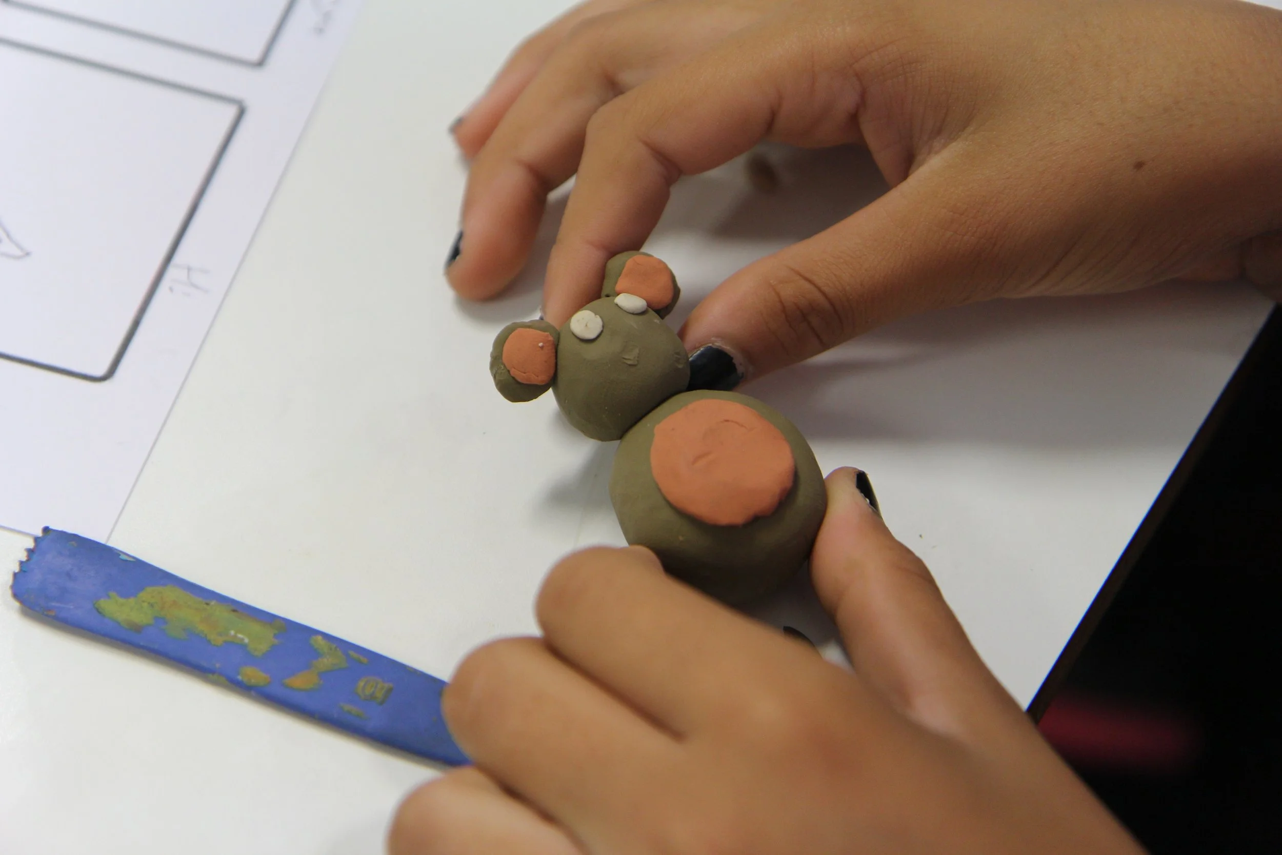 Person's hands sculpting a small, cute bear figure with clay on a white surface, with a paint stir stick nearby.