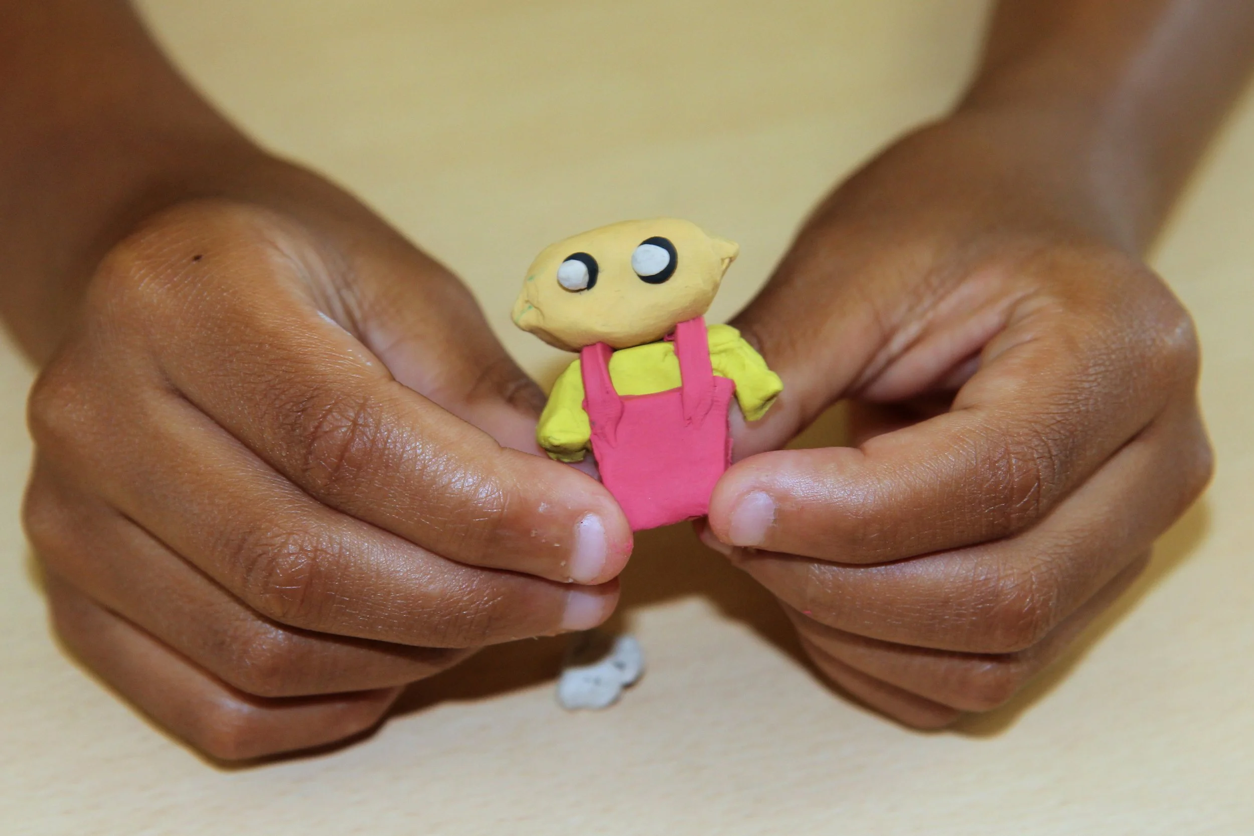 Hands holding a small handmade clay figure of a character with a yellow head, large eyes, yellow shirt, and pink overalls.