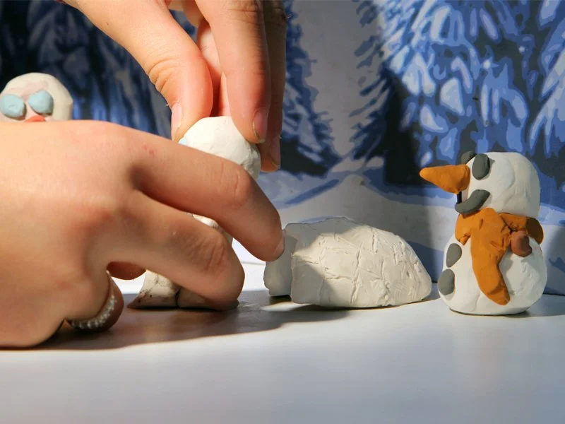 Hands shaping a snowman out of white modeling clay next to other clay snowman figures on a table.