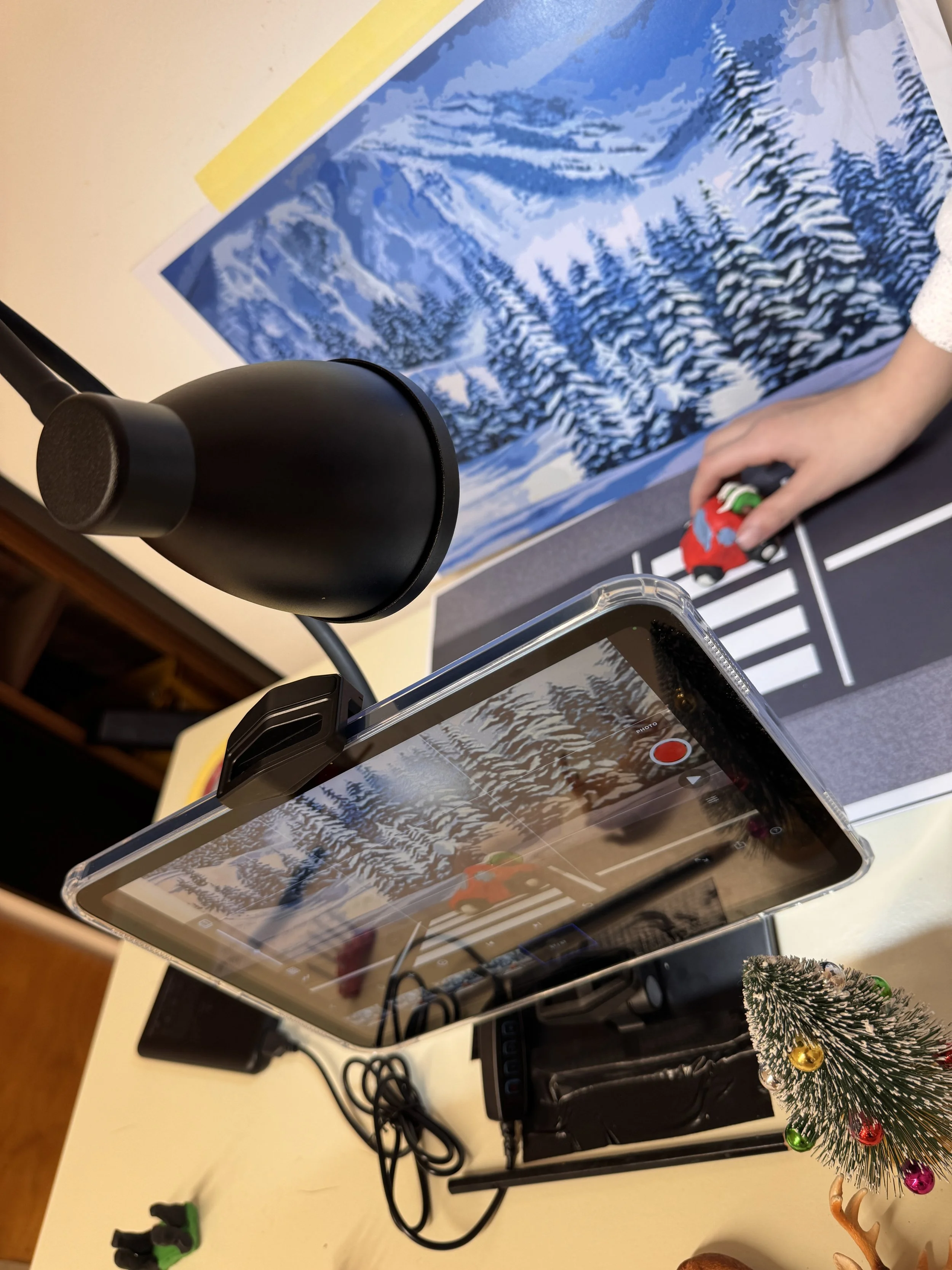 A tabletop setup for filming a stop-motion animation with a winter landscape illustration, a smartphone on a stand, a small Christmas tree, and a person holding a toy car on a striped background.
