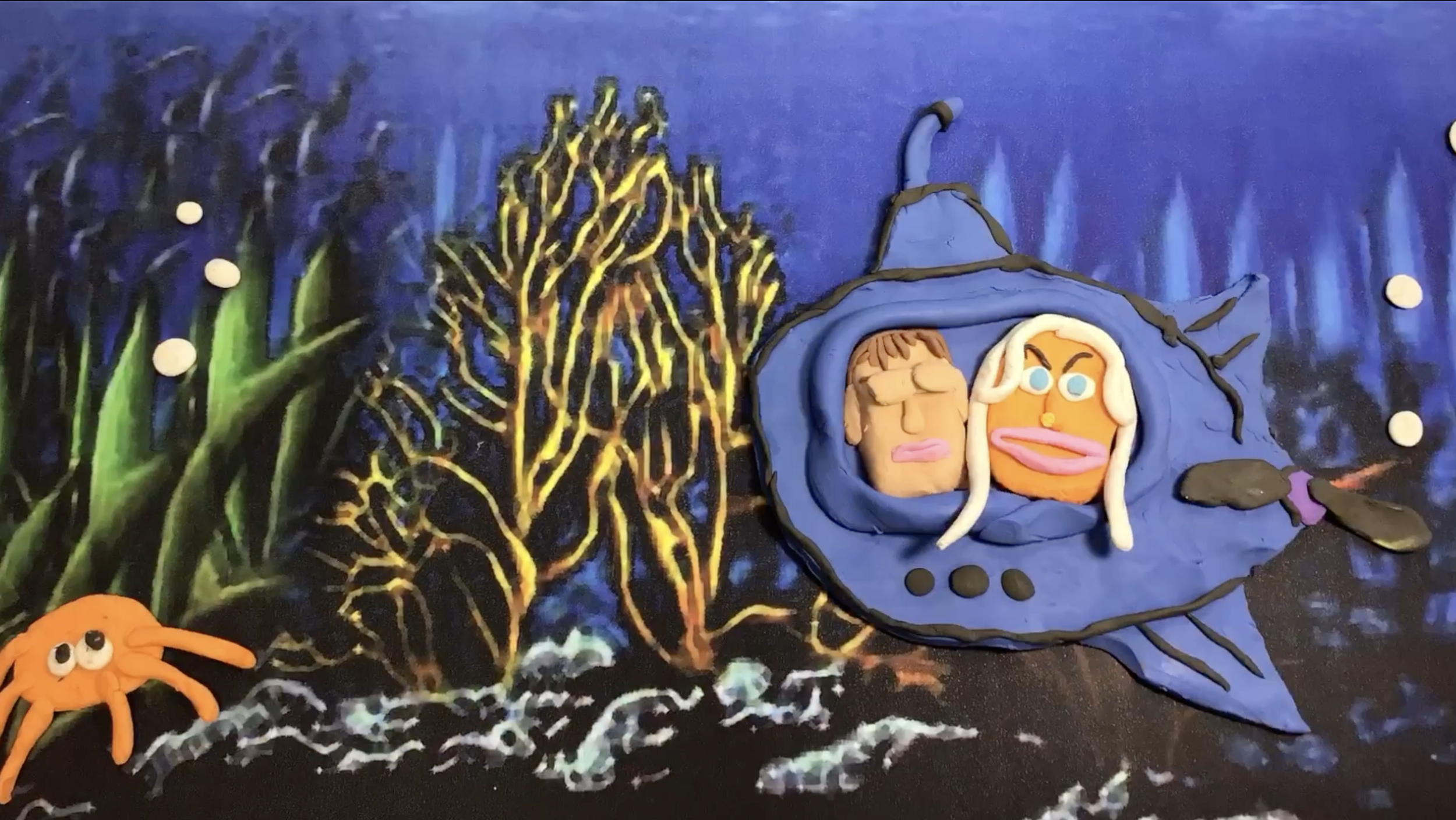 Crafted clay scene of an underwater ocean with coral, a small orange octopus, and a large blue fish with human-like face features and blonde hair, set against a dark underwater background with bubbles.