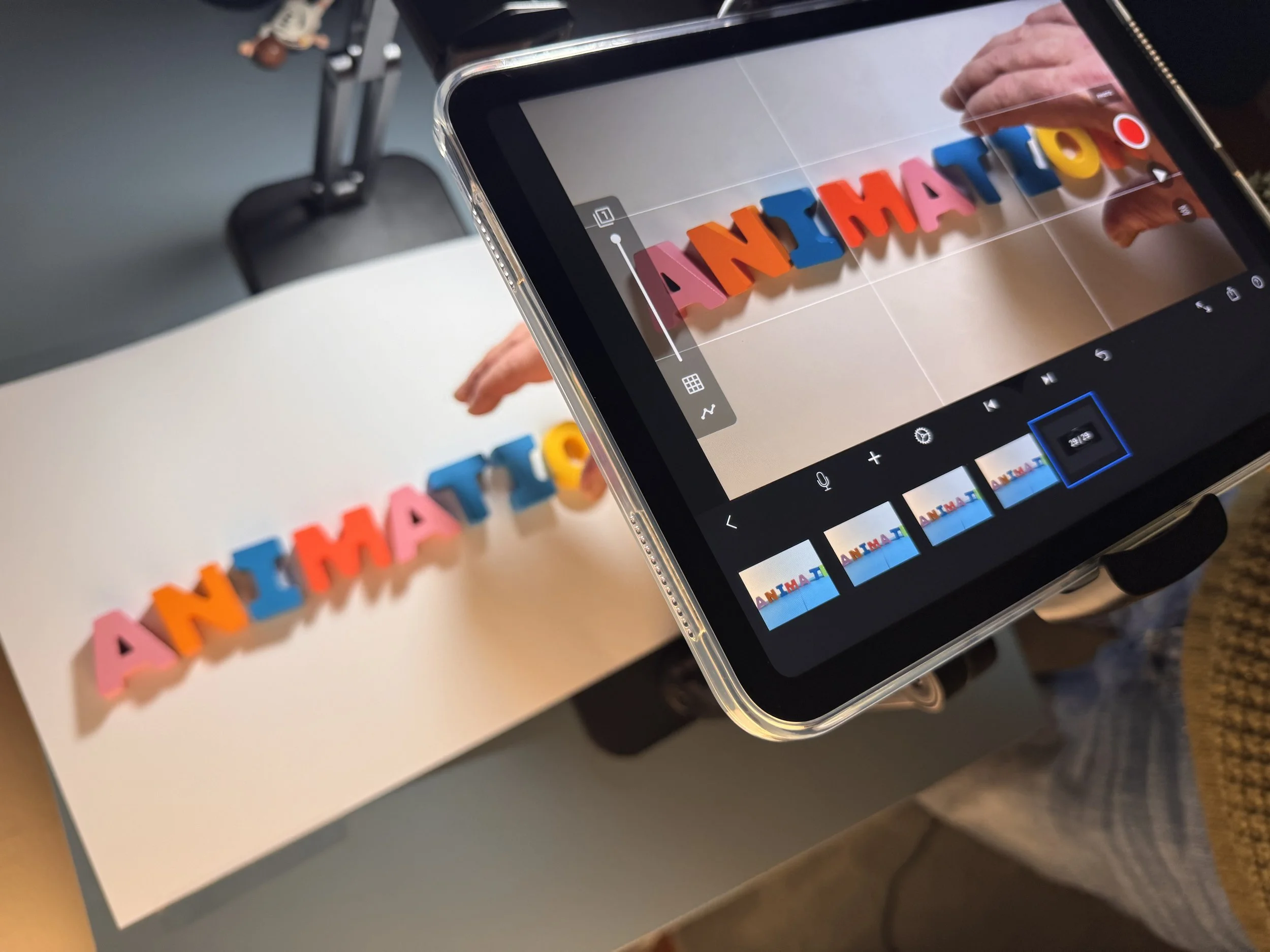A person is taking a photo of colorful letter toys spelling 'ANIMATION' on a white surface with a smartphone. The phone screen shows the subject through the camera.